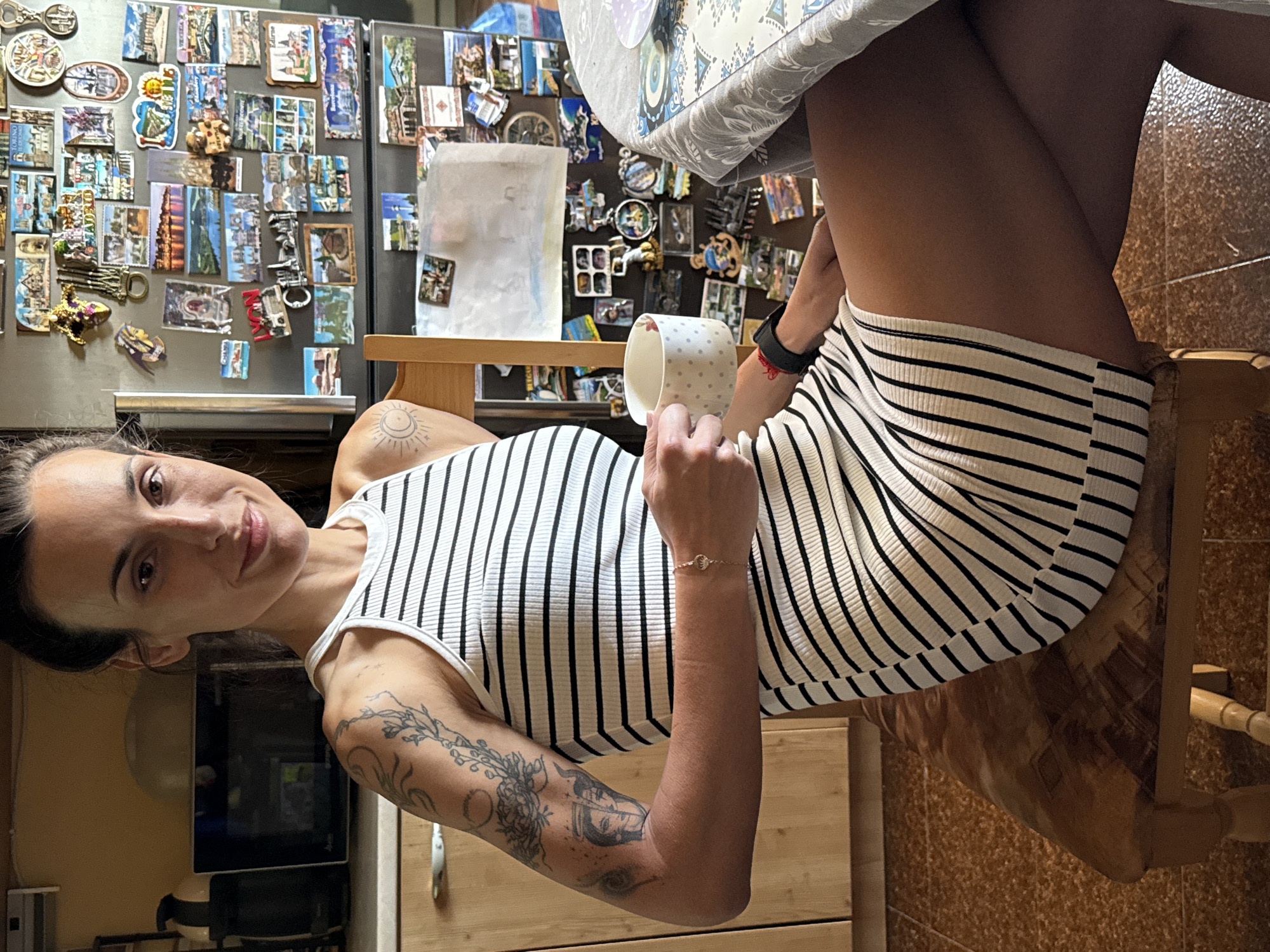 The media file displays an indoor scene, rotated 90 degrees clockwise from its natural orientation. It features a young woman seated at a wooden table, likely in a kitchen or dining area in Pleven, Bulgaria. She is looking towards the viewer with a gentle smile, holding a white mug with grey polka dots in her right hand.
The woman has dark hair pulled back and is wearing a white tank top or dress with thin black horizontal stripes. Visible tattoos adorn both of her arms: a sun and crescent moon design is on her left shoulder, and intricate floral and abstract patterns cover her left forearm. A delicate gold-colored bracelet is on her right wrist.
Behind her, a stainless steel refrigerator is heavily decorated with numerous souvenir magnets from various locations. Visible text on some magnets includes "MADRID", "TORINO", "ROMA", "BARCELONA", "VICTORIA", "NEW YORK", and "PARIS". A white paper note is also affixed to the refrigerator. To the left of the woman, a dark-colored appliance, possibly a microwave or oven, is visible on a wooden surface. The table she sits at is covered with a patterned tablecloth featuring blue and grey geometric designs. The overall lighting suggests an indoor artificial light source, contributing to a relaxed atmosphere.