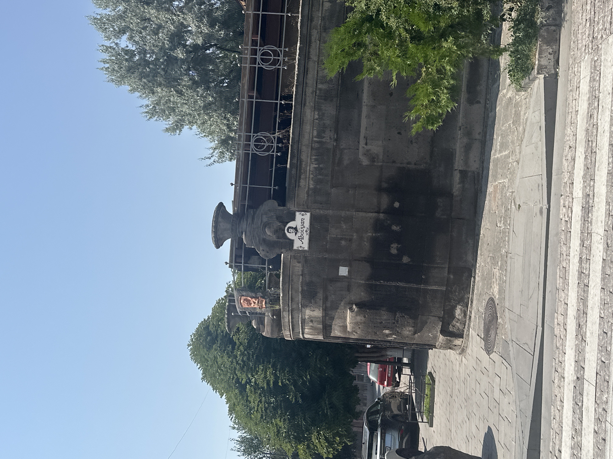 The image shows a street scene in Gyumri, Armenia, on a clear, sunny day. In the foreground, a pedestrian crossing with white stripes is visible on a paved road. To the left, a gray car is parked. Behind the car, a red car is also parked.
The dominant feature of the scene is a large, dark stone structure that appears to be part of a building or monument. This structure has a rounded corner and is decorated with urns and ornate railings. A sign with a portrait and the name "Abovyan" is affixed to the stone. Behind the stone structure, a large, leafy green tree stands tall. To the right, another green tree with feathery foliage is visible. The sky above is a clear, bright blue. The overall impression is of a well-maintained urban environment, likely a public square or street with historical elements.