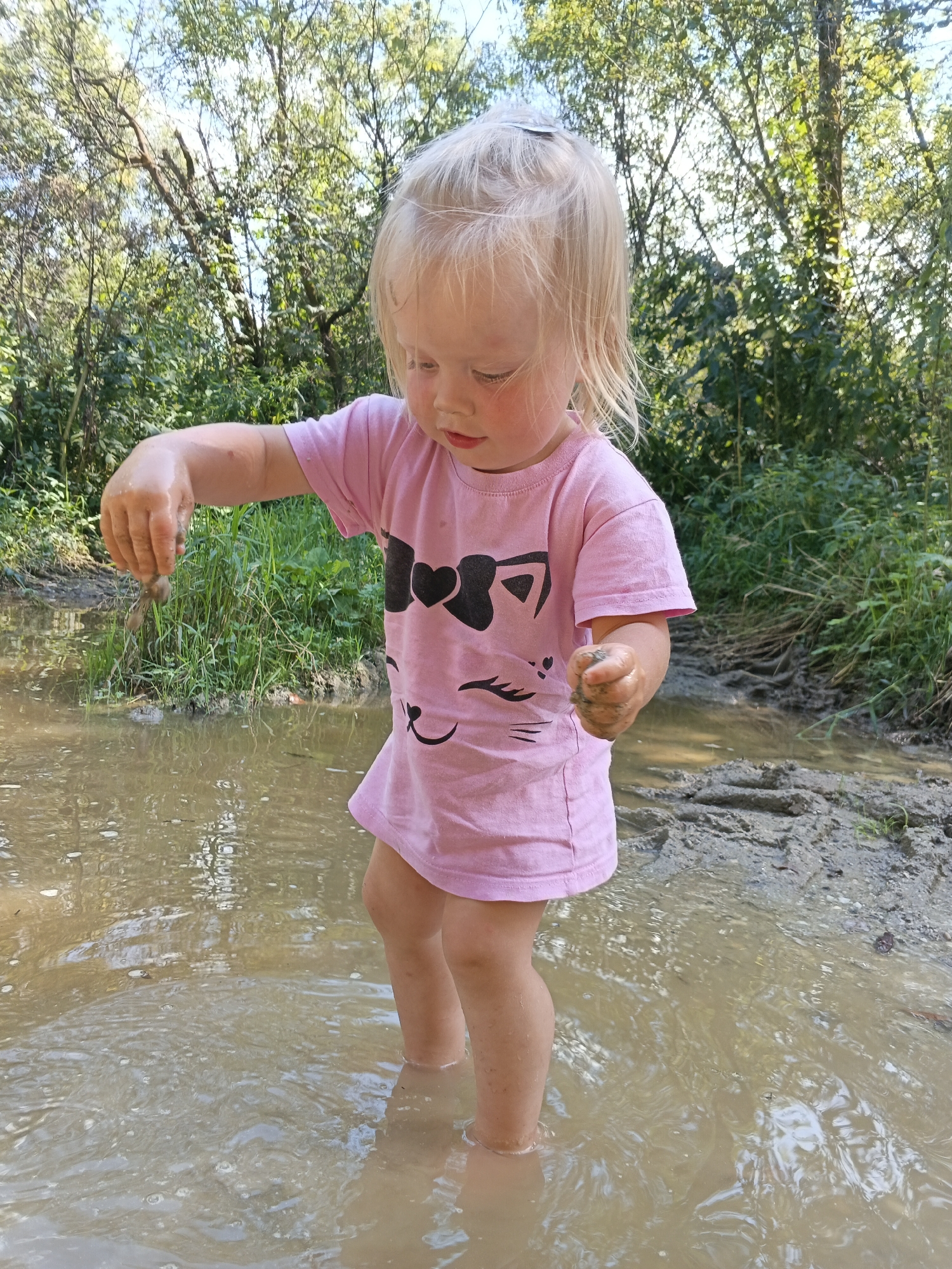 A young child with blonde hair is standing in shallow, muddy water, holding a small clump of dirt in each hand. The child is wearing a pink t-shirt with a black cat face and bow design. The surrounding environment is natural, with trees and green foliage visible in the background. The ground is muddy and wet, indicating a possible stream or puddle. The lighting suggests it is daytime. The location is specified as Medvedyovka, Russia.