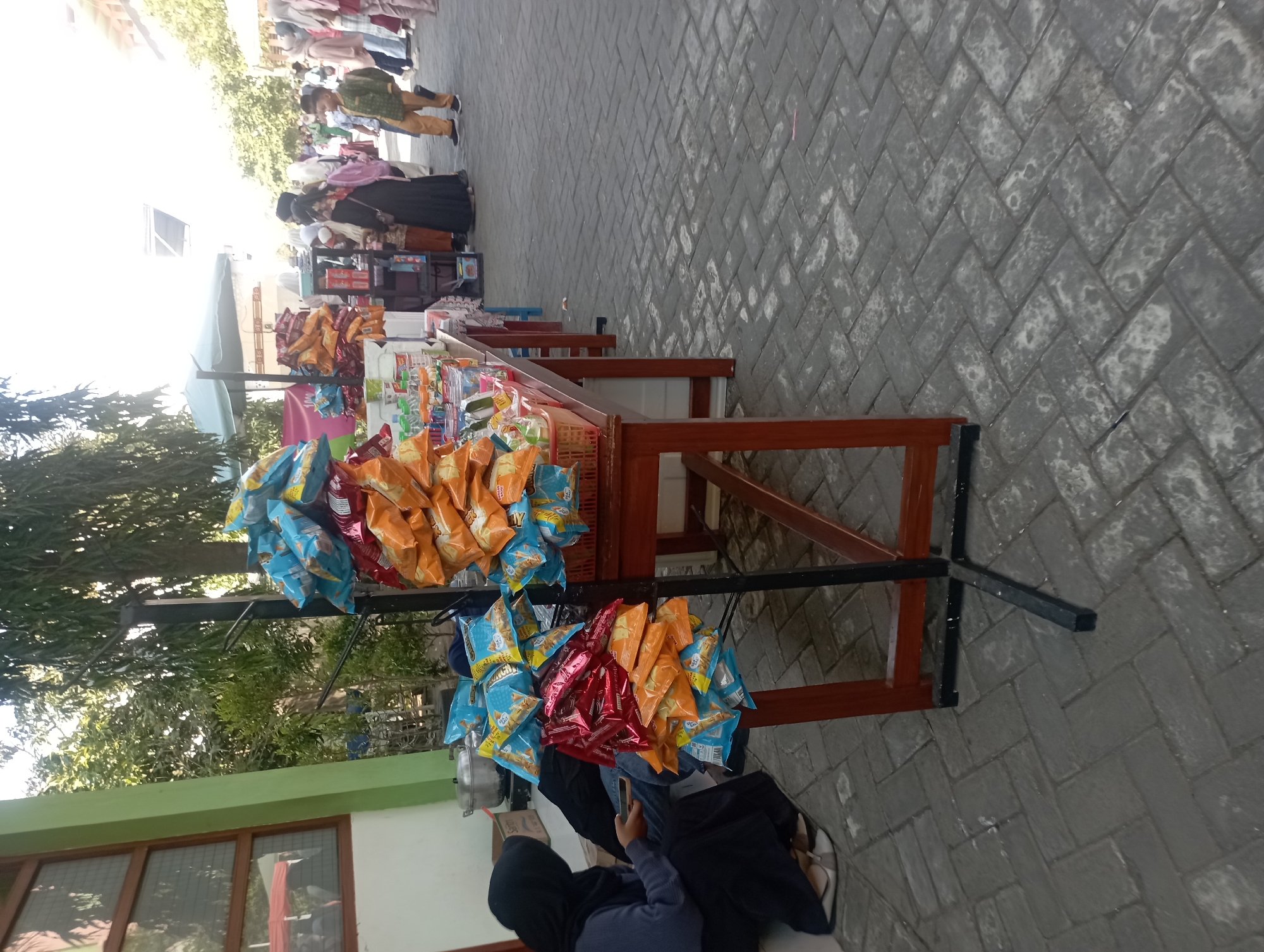 The image depicts a street-side stall selling snacks in Nganjuk, Indonesia. The stall is set up along a brick-paved walkway, with a building visible to the left and trees providing shade. Several people are browsing or working at the stalls, including a woman in a black hijab sitting near the foreground and a young boy in a green outfit standing by another stall further down.
The main focus of the image is the display of snacks, predominantly in colorful plastic packaging. Bags of chips and other processed food items are arranged on a rack and on tables. Some tables also hold bottles of drinks and smaller packaged goods. The stalls are simple, made of wood and cloth awnings, suggesting a temporary or informal setup. The overall atmosphere appears to be that of a local market or event where vendors are selling refreshments. The time of day seems to be daytime, given the sunlight and shadows.