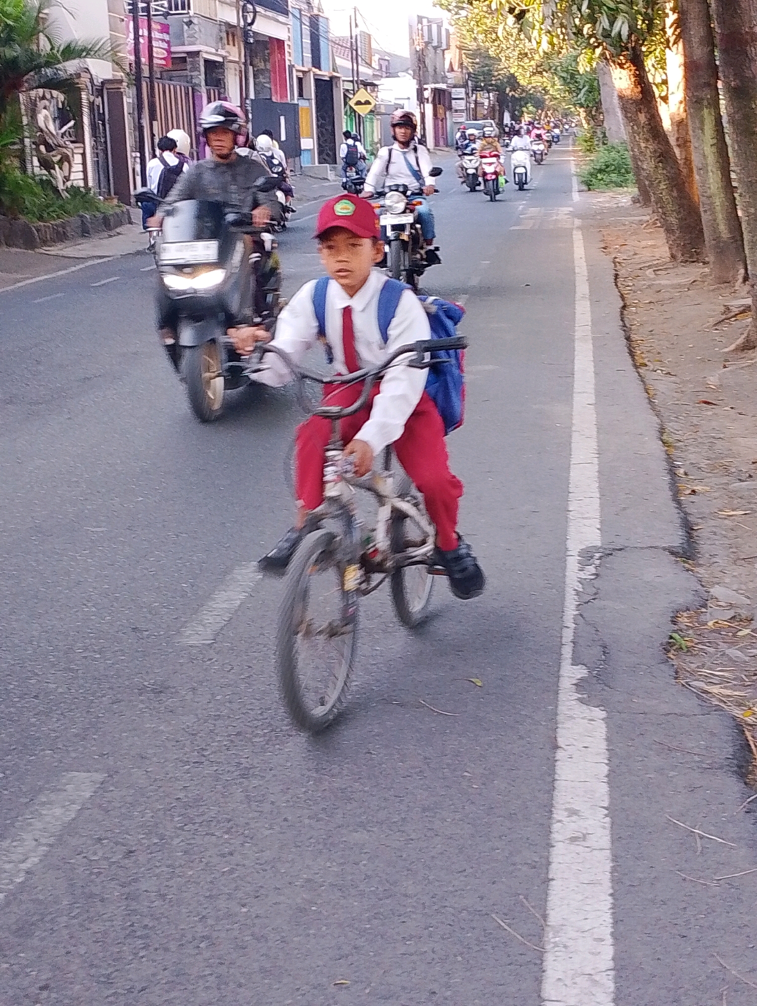 The image depicts a street scene in Probolinggo, Indonesia, with a young boy in the foreground riding a bicycle. He is wearing a school uniform consisting of a white shirt, red pants, and a red cap with a green emblem. A blue backpack is slung over his shoulders. The boy is in motion, as indicated by the blur of his bicycle's wheels. Behind him, several motorcycles are also in motion, suggesting a busy street. The motorcycles are diverse, with riders wearing helmets. The environment appears to be a city street with buildings lining one side and trees on the other. The lighting suggests it is either early morning or late afternoon, with sunlight filtering through the trees.