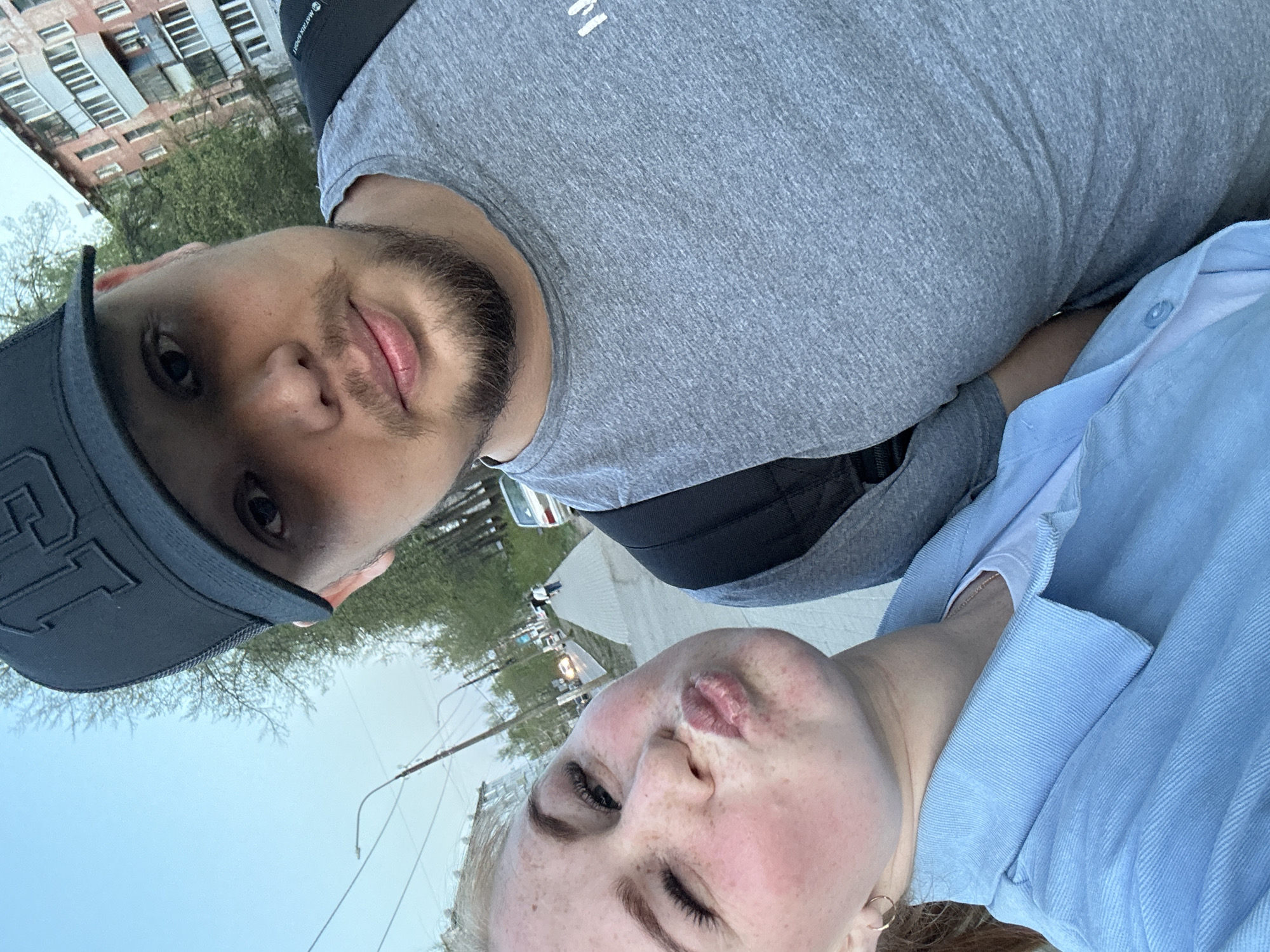 A selfie taken outdoors in daylight shows two people. A person with freckles is at the bottom of the frame with pursed lips and one eye closed. They wear a light blue shirt and a small hoop earring. A person wearing a gray t-shirt and a dark cap is at the top of the frame. This person has a beard. In the background are buildings and trees along a city street with street lights. Based on the buildings and street layout, the location is likely Chaikovsky, Russia.