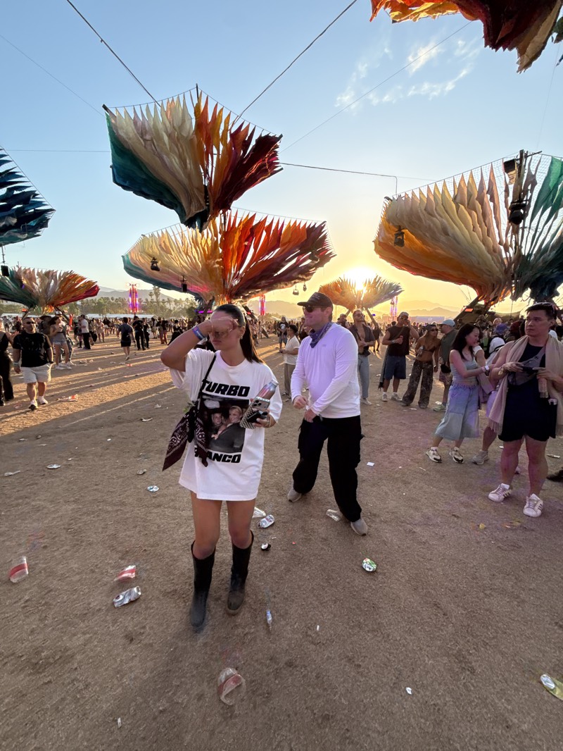 The image shows an outdoor scene at a festival in the city of Indio, California, United States. The ground is dry and littered with trash, specifically crushed cans and other debris. There are multiple people in the background, appearing to be festival attendees, with a mix of clothing styles. Above the crowd, there are large, colorful, decorative structures resembling stylized leaves or petals suspended on wires, creating an artistic canopy.
In the foreground, there is a woman wearing a white oversized T-shirt with a graphic print, short black boots, and a headband. She has sunglasses on and is holding one hand to her head. Next to her, a man is wearing a white long-sleeved shirt, a baseball cap, and dark trousers. He appears to be in motion or dancing. The setting sun is visible in the background, providing a warm light to the scene.