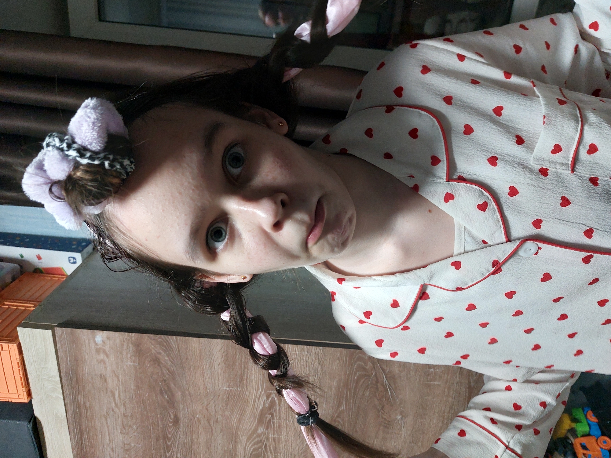 The image shows a young person indoors in Chaikovsky, Russia, looking directly into the camera with a wide-eyed and slightly open-mouthed expression that suggests surprise or playfulness. Her dark hair is styled in two distinct ways: on the right side of her head (viewer's left), a fluffy light purple scrunchie holds a bun, while on the left side of her head (viewer's right), a long braid is visible, interwoven with a light pink ribbon and secured at the end with a black hair tie. A small gold earring is visible in her left ear.
She is wearing a light-colored top, likely pajamas or loungewear, patterned with numerous small red heart shapes. The top features red piping along its collar and front opening, with a white button visible near her right shoulder. Her complexion appears light, with some minor blemishes or freckles.
The background indicates an informal home setting. A wooden cabinet or dresser with a dark top surface occupies the lower half of the frame. To the left, orange plastic crates are stacked on a surface, with a blue and white box partially visible above them. To the lower right, colorful plastic toys are visible. A window or dark frame is in the upper background, providing an indoor context. The lighting suggests it is daytime. No text is visible in the image.