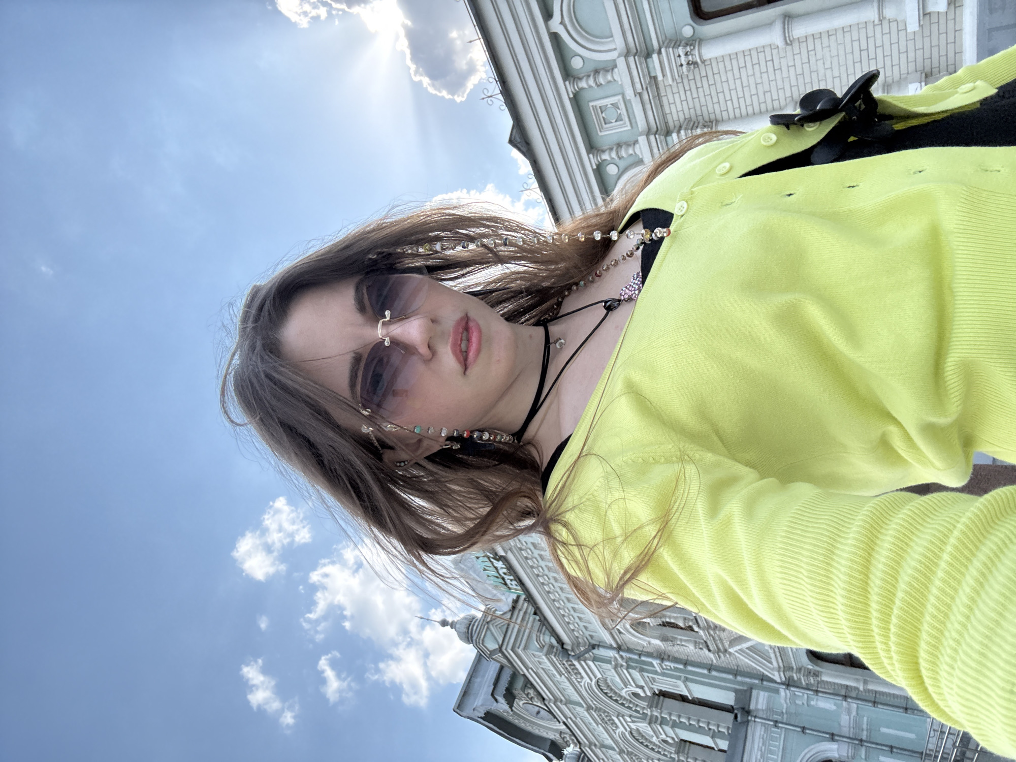 A young woman stands in front of an elaborate, light-colored building in Moscow, Russia, possibly the GUM department store. She wears sunglasses, a black choker, a bright yellow cardigan, and beaded strands in her hair. The sky is blue with scattered clouds, and the sun is shining. The camera angle is from below, looking up at the woman and the building.