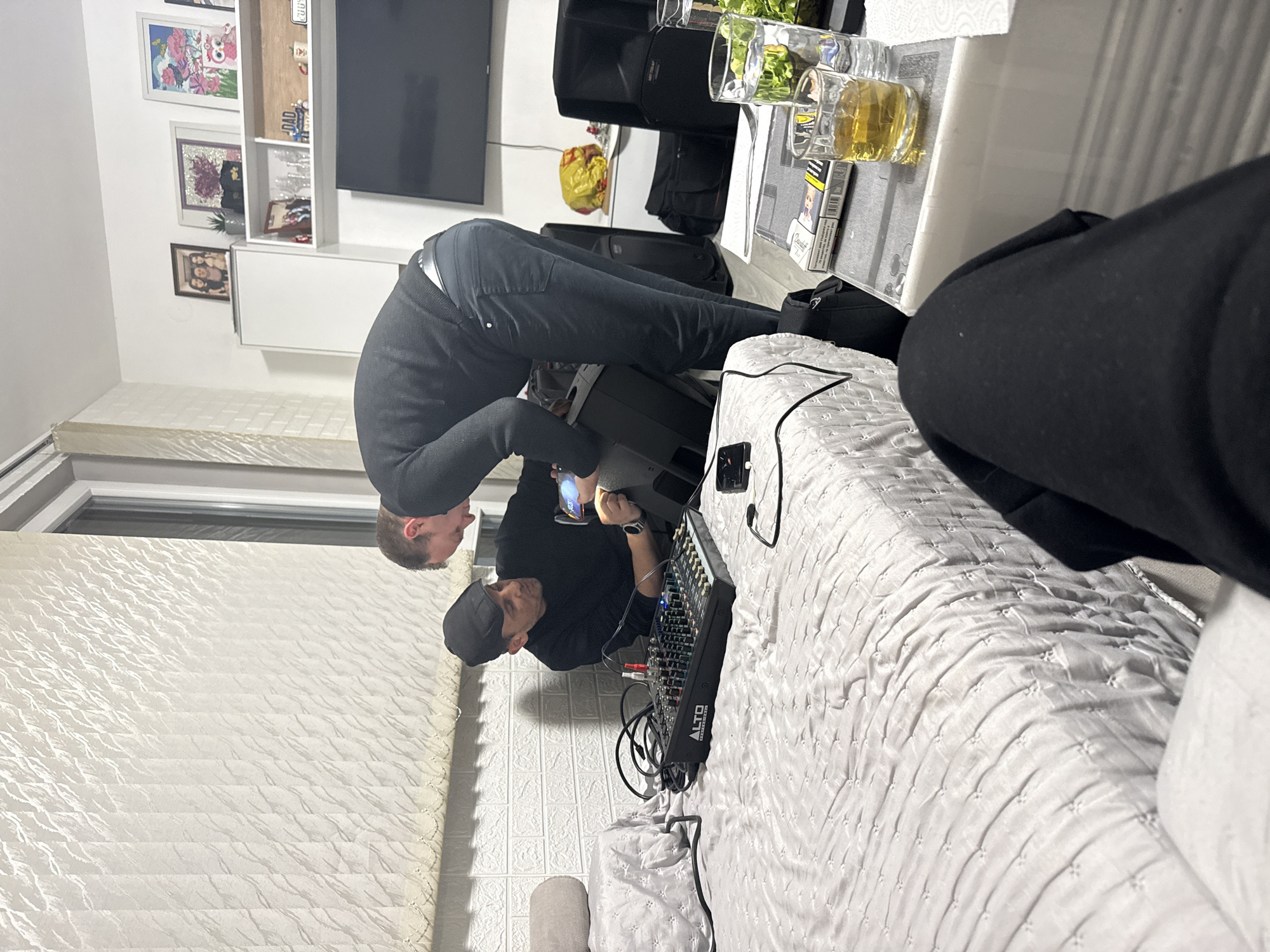 Two men are indoors. One man stands, leaning over a piece of equipment. He wears black clothing. The second man crouches, looking at a cell phone, also wearing black clothing and a cap. There is sound mixing equipment on a bed. The room has a television mounted on the wall, shelving and family photos. The location appears to be a home, not in Sofia, Bulgaria.
