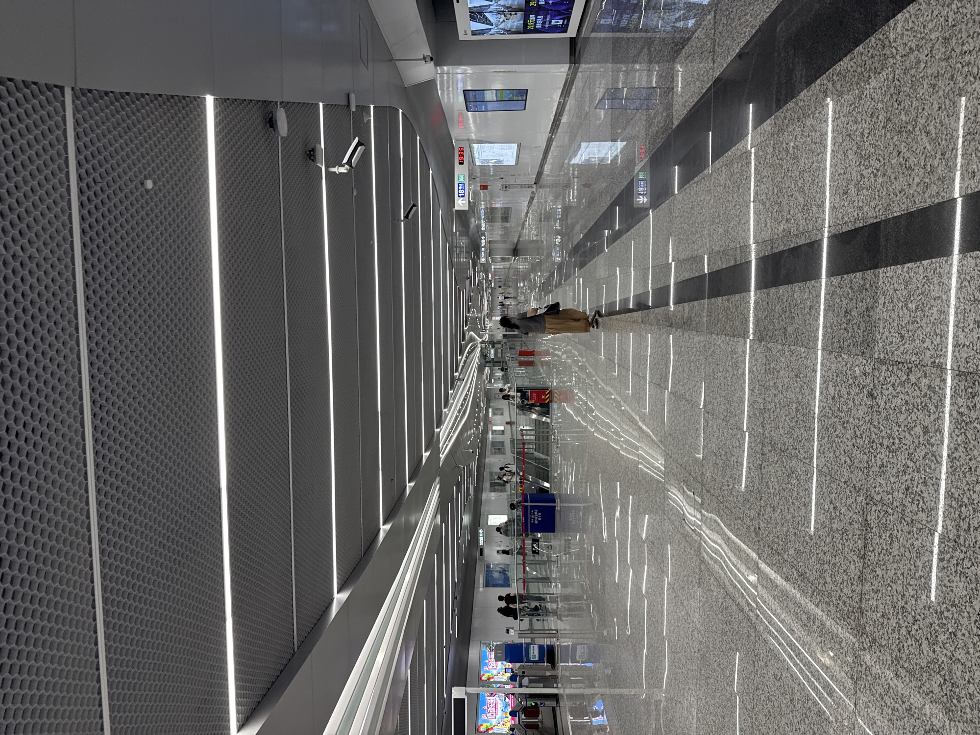 The image depicts a modern, brightly lit subway station in Tianhe District, China. The perspective is from a high vantage point, looking down a long, straight platform. The walls on the left are adorned with a textured, honeycomb-like pattern, punctuated by vertical LED strip lights that create a sleek, futuristic ambiance. On the right, the floor is a polished granite, also marked by parallel lines of embedded lighting that reflect the illumination.
Several individuals are visible, scattered along the platform. One person in the foreground is facing away from the camera, seated with their belongings. Further down the platform, other passengers are standing, some near escalators or entrances, suggesting they are either waiting for a train or navigating the station. Security cameras are mounted on the walls.
Signs with Chinese characters are visible, including directional arrows and station information, though the text is not fully legible due to the distance and resolution. One sign displays a subway map or route information. The overall scene suggests a typical day in a busy urban transit hub. The lighting is bright and consistent, implying indoor illumination rather than natural daylight, and there are no obvious weather-related cues. The atmosphere appears orderly and functional.