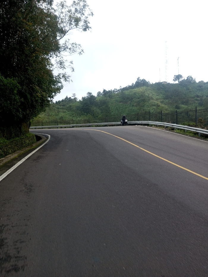 A winding asphalt road with a solid yellow line running down the center curves through a hilly landscape. A metal guardrail lines the road's edge on one side. Trees and foliage border the other side of the road. A lone motorcyclist sits on the side of the road. Towers are visible in the distance atop a grassy hill. The sky is overcast.