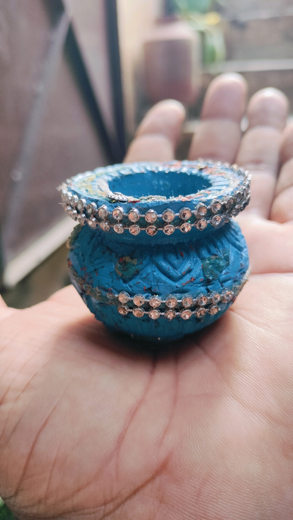 A close-up, low-angle shot captures a small, blue, decorative pot held in the palm of a person's hand. The pot, made of what appears to be clay, is adorned with two horizontal bands of small, clear rhinestones around its rim and lower body. The blue paint is textured and shows some red and green embellishments. The hand, which is light-skinned, is cupped to support the pot, with visible palm lines and knuckles. The background is blurred, suggesting an outdoor or semi-outdoor environment with hints of greenery and structures. The setting is identified as Ramraj, India.