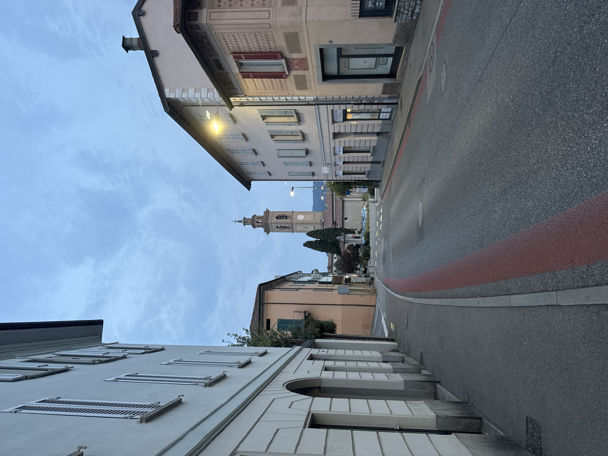 The image depicts a street scene in Balerna, Switzerland, at dusk. The perspective is from street level, looking up and along a gently curving road that leads towards a distant bell tower. On the left, buildings line the street, characterized by their muted pastel colors and architectural details such as decorative friezes and numerous windows with closed shutters. The building closest to the viewer is a light-colored structure with a patterned facade. Further down the street, a taller, light pink building is visible, followed by a peach-colored building with green shutters. The road itself is asphalt, with a distinct red and gray stripe marking a lane or pathway. In the distance, the bell tower of a church rises above the buildings, its clock face visible. The sky is overcast with scattered clouds, and artificial lights from street lamps and buildings illuminate the scene. The overall atmosphere is quiet and serene, suggesting the end of the day. No people are visible in the image.