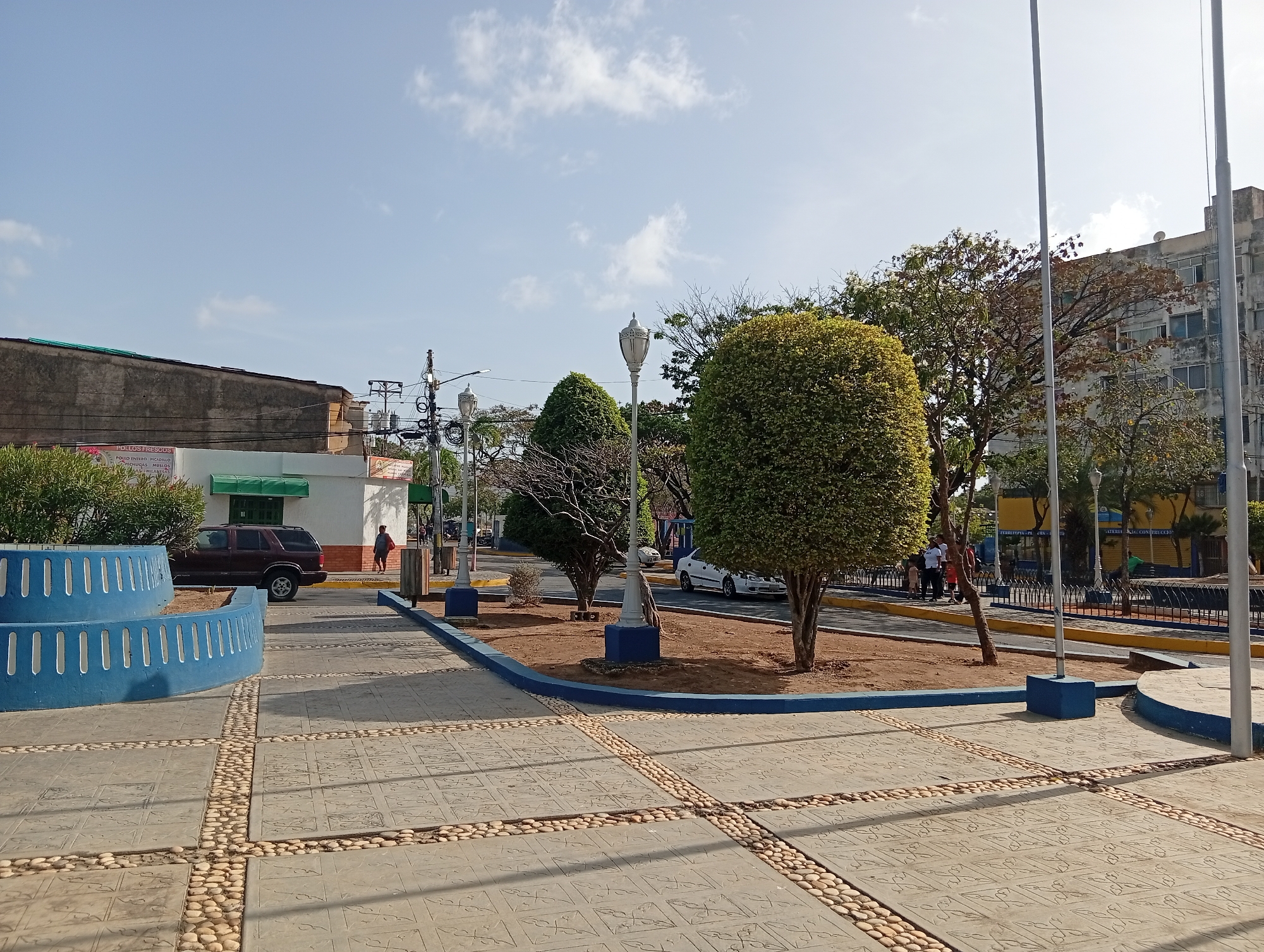 The image depicts a public square in Juan Griego, Venezuela, on a clear day. The square is paved with square concrete slabs separated by rows of small cobblestones. In the foreground, there is a blue, tiered, semi-circular structure with vertical openings. The ground is patterned with geometric designs.
Central to the scene are three trees with rounded canopies, planted in a bed of red soil bordered with a blue curb. There are ornate, vintage-style lamp posts with white globes and blue bases situated among the trees. Behind the trees, a street is visible with parked cars and pedestrians. In the background, buildings of varying heights line the street, displaying a mix of architectural styles.
A dark-colored SUV is parked to the left of the frame. A person is walking on the sidewalk behind the SUV, and other individuals are visible further down the street.
The sky is pale blue with scattered, light clouds. The scene is bathed in sunlight, casting shadows on the pavement. Two tall flagpoles stand on the right side of the image.