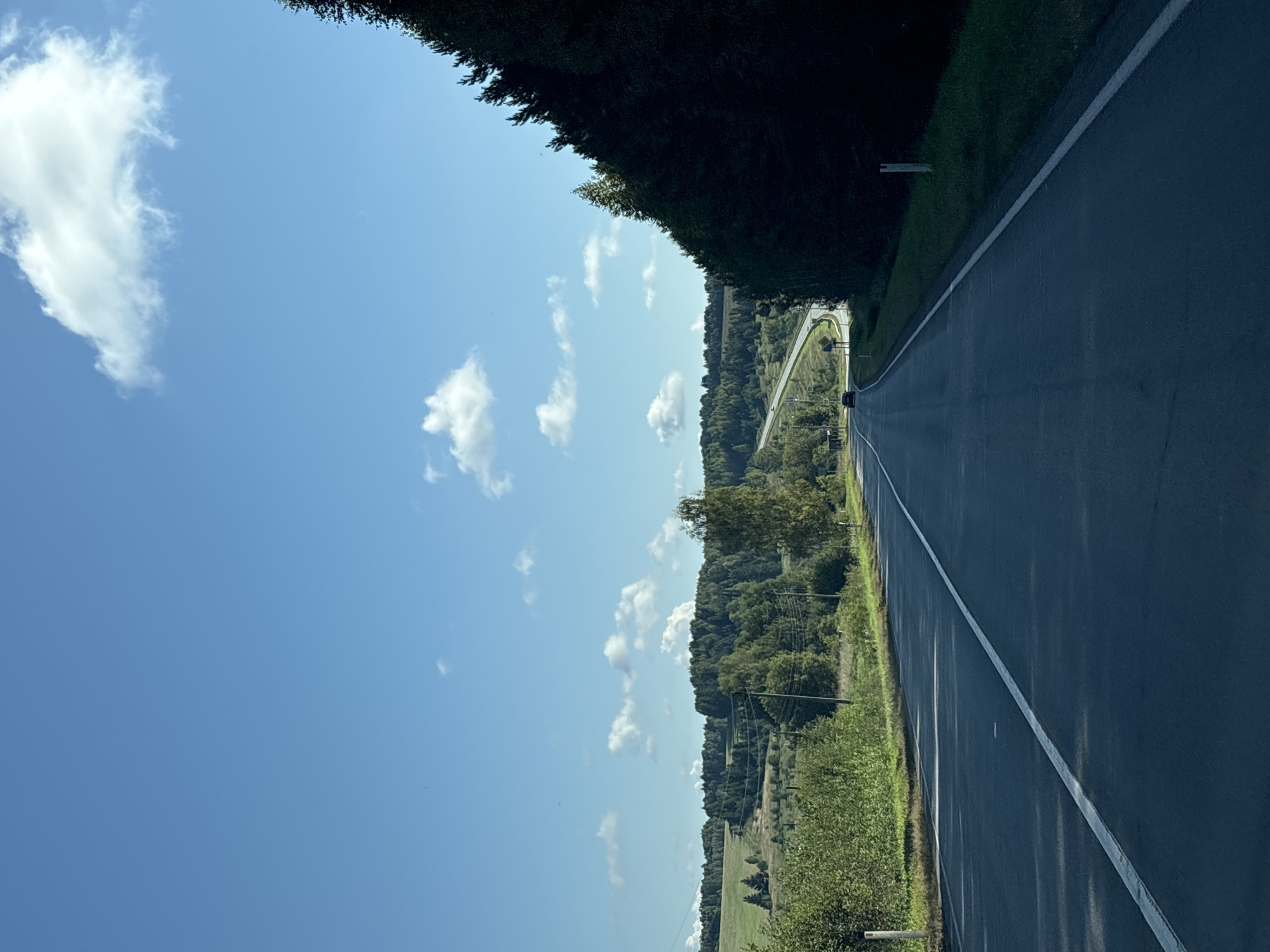 A straight, paved road with a white line dividing the lanes stretches into the distance under a clear blue sky dotted with white clouds. The road curves gently to the left as it ascends a hill. On the right side of the road, a dense line of dark green trees forms a shadow, while on the left, a green field slopes upwards, dotted with scattered trees. In the far distance, a few buildings and more trees are visible, suggesting a rural or suburban setting. A dark-colored car is traveling on the road in the distance. The overall scene depicts a peaceful, scenic drive in a rural landscape.