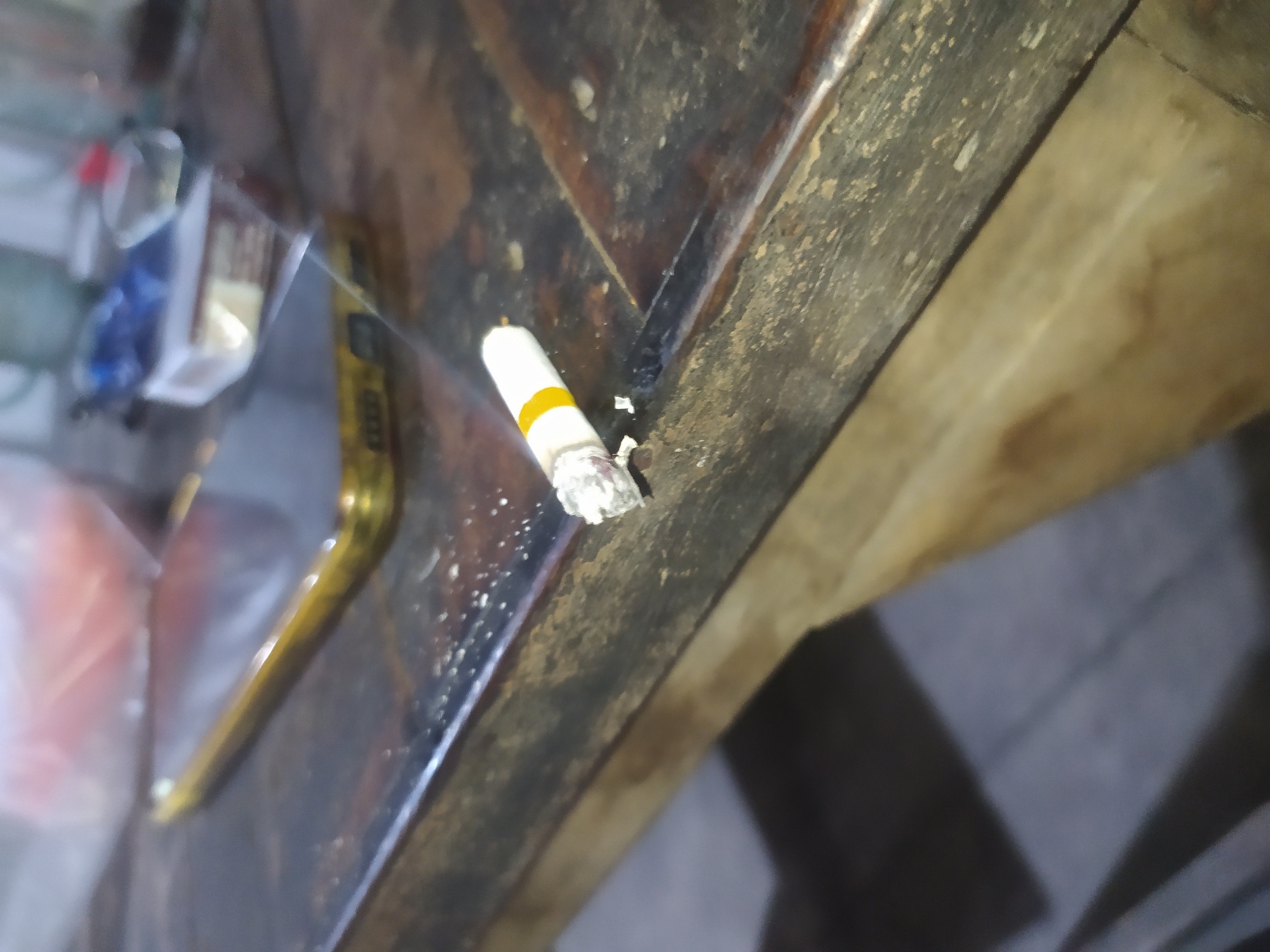 The image shows a close-up of a lit cigarette resting on a dark wooden table. The cigarette is partially burned and appears to be made of tobacco with a white filter and a yellow band near the tip. The table surface is rough and worn, with visible imperfections and a slight sheen. In the background, a gold-colored smartphone is visible, along with a pack of cigarettes and other indistinct objects that appear blurred and out of focus. The lighting suggests an indoor environment, possibly dim. There are no people visible in the image, and no specific details about the city of Tuntang, Indonesia, can be inferred from the scene.