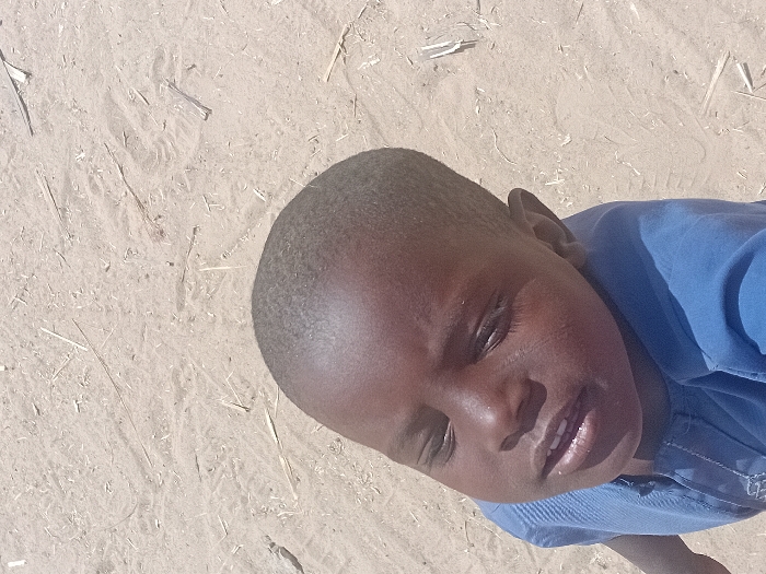 A young child, located in Ayama, Nigeria, is depicted from a high-angle, looking upwards towards the viewer. The child presents with dark skin, short dark hair, and is wearing a blue short-sleeved garment with vertical textured stitching on the front and a faint light-colored design near the neckline. The child's eyes are partially closed, appearing to squint, and the mouth is slightly open, revealing some teeth. The background consists of light tan-colored sand, marked with scattered fragments of dry organic material, such as twigs and straw, along with faint surface impressions. The scene is brightly illuminated, suggesting direct overhead sunlight.