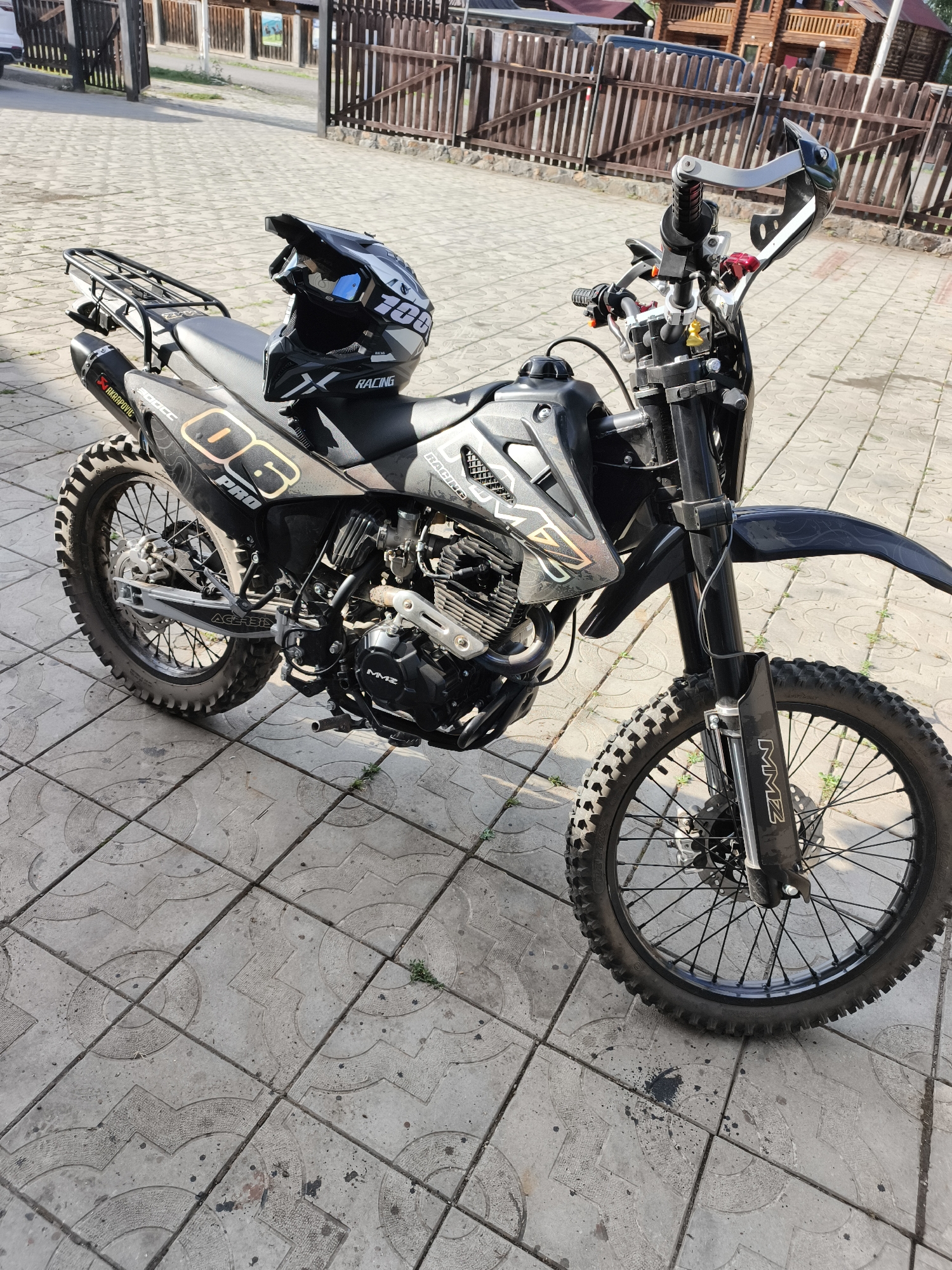 A black dirt bike with a silver and black graphic design is parked on a paved patio. The motorcycle is facing towards the right of the frame. A black helmet with silver and white accents sits on the seat of the motorcycle. The background shows a wooden fence and a wooden building. The ground is paved with grey concrete tiles that have a decorative pattern.