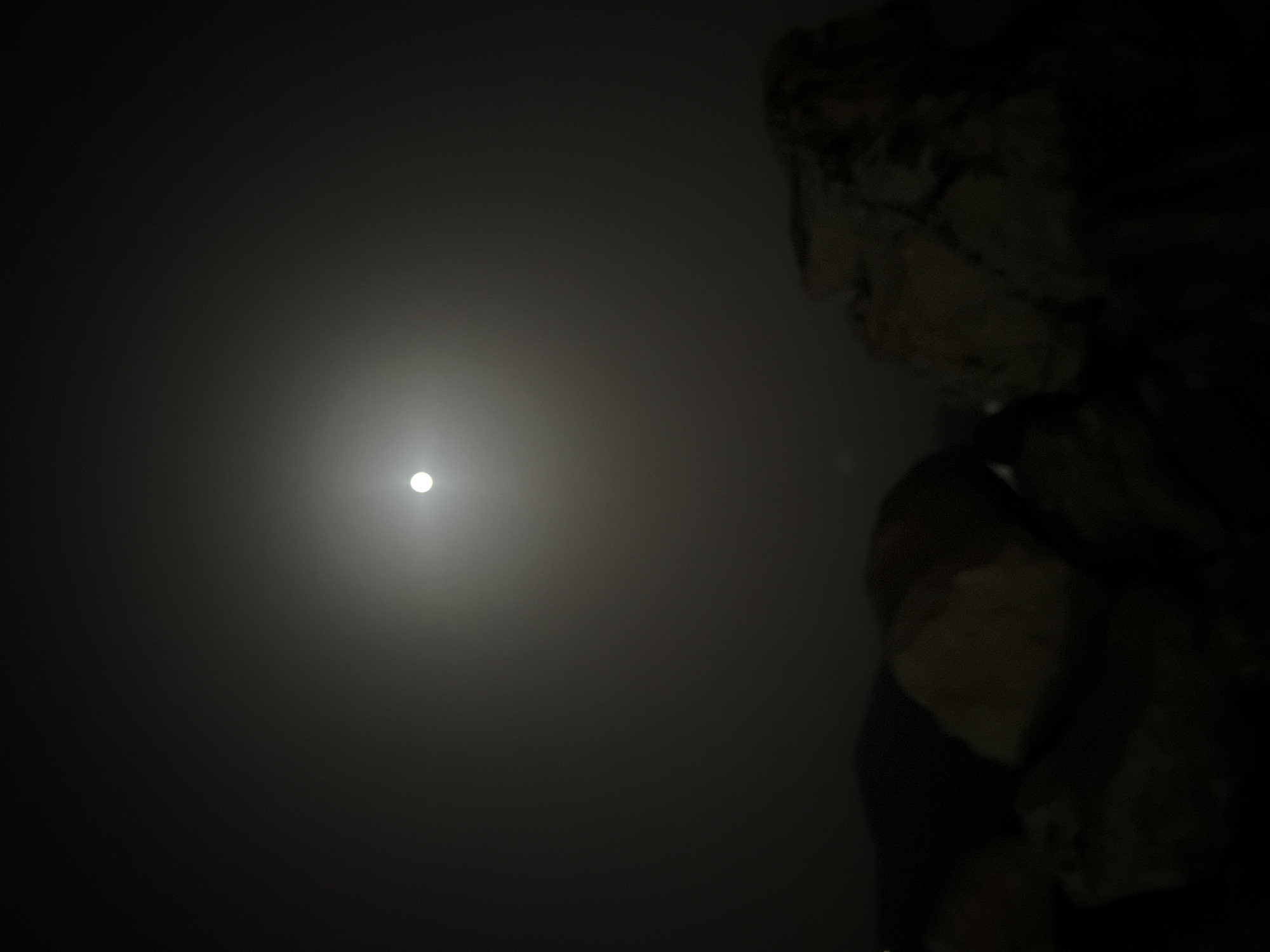 The image depicts a nighttime scene with a full moon in a hazy sky. The moon is a bright, white orb positioned in the upper center of the frame, surrounded by a soft glow, suggesting fog or atmospheric moisture. In the foreground, the silhouettes of large rocks are visible, some appearing light-colored and others darker, indicating they are illuminated by the moonlight. The overall setting appears to be an outdoor, possibly coastal, environment given the presence of rocks. There is no indication of the specific location within the Municipal Unit of Alyzia, Greece, or any human presence.