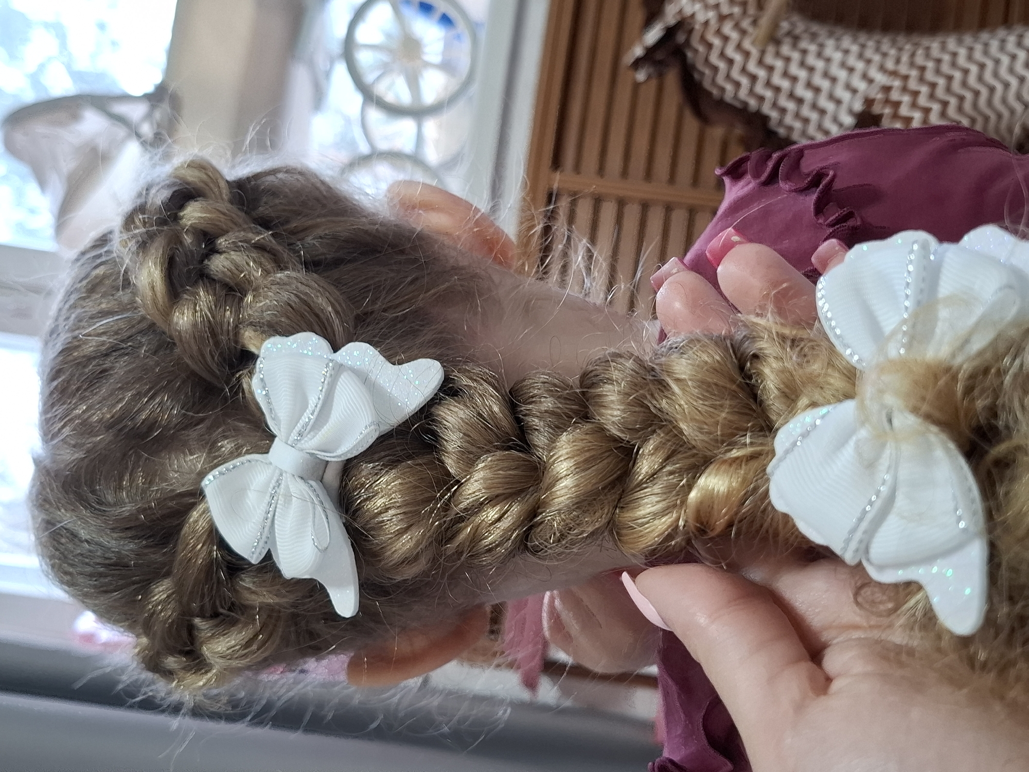 A close-up shows the back of a doll's head with light brown hair styled in two braids, each adorned with a white bow featuring a glittery trim. A fair-skinned hand with polished nails is partially visible, seemingly holding the doll's head. The doll is wearing a garment of dark pinkish-red fabric. The background consists of a window, part of a wicker object, and a brown and white patterned object. Location: Chaikovsky, Russia.