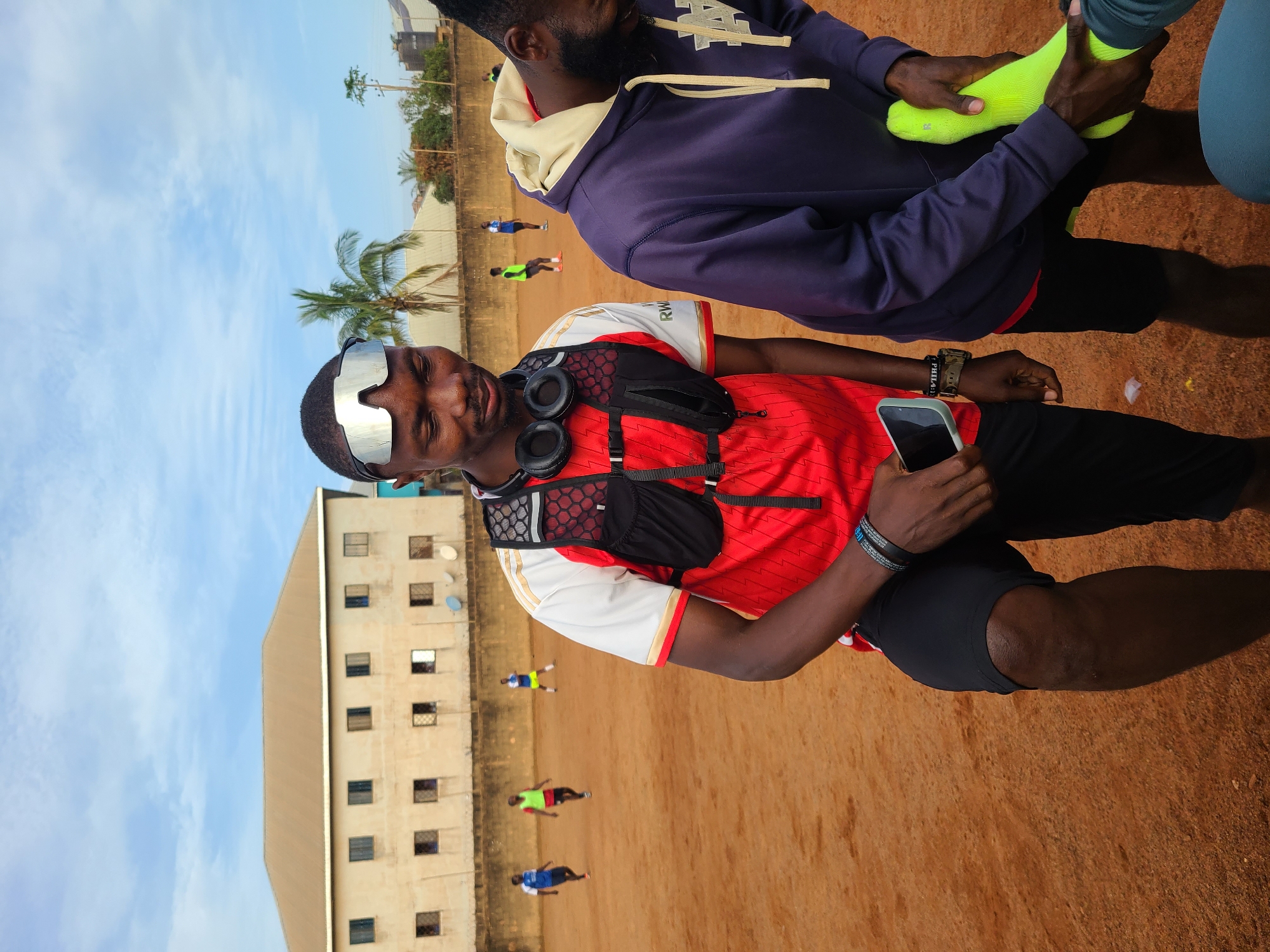 A group of young men are on a dusty, reddish-brown outdoor soccer field in Barakin Lerawi, Nigeria. In the foreground, two men are facing each other. The man on the left, wearing a red and white jersey, black shorts, a tactical vest, and silver-tinted goggles, is holding a smartphone. The man on the right, in a purple hoodie, is putting on bright yellow soccer socks. In the background, several other men are scattered on the field, some appearing to be playing soccer. Behind the field, a low concrete wall is visible, topped with a brick wall. Beyond that, a long, beige, two-story building with multiple windows can be seen, along with palm trees and a water tank. The sky is partly cloudy.