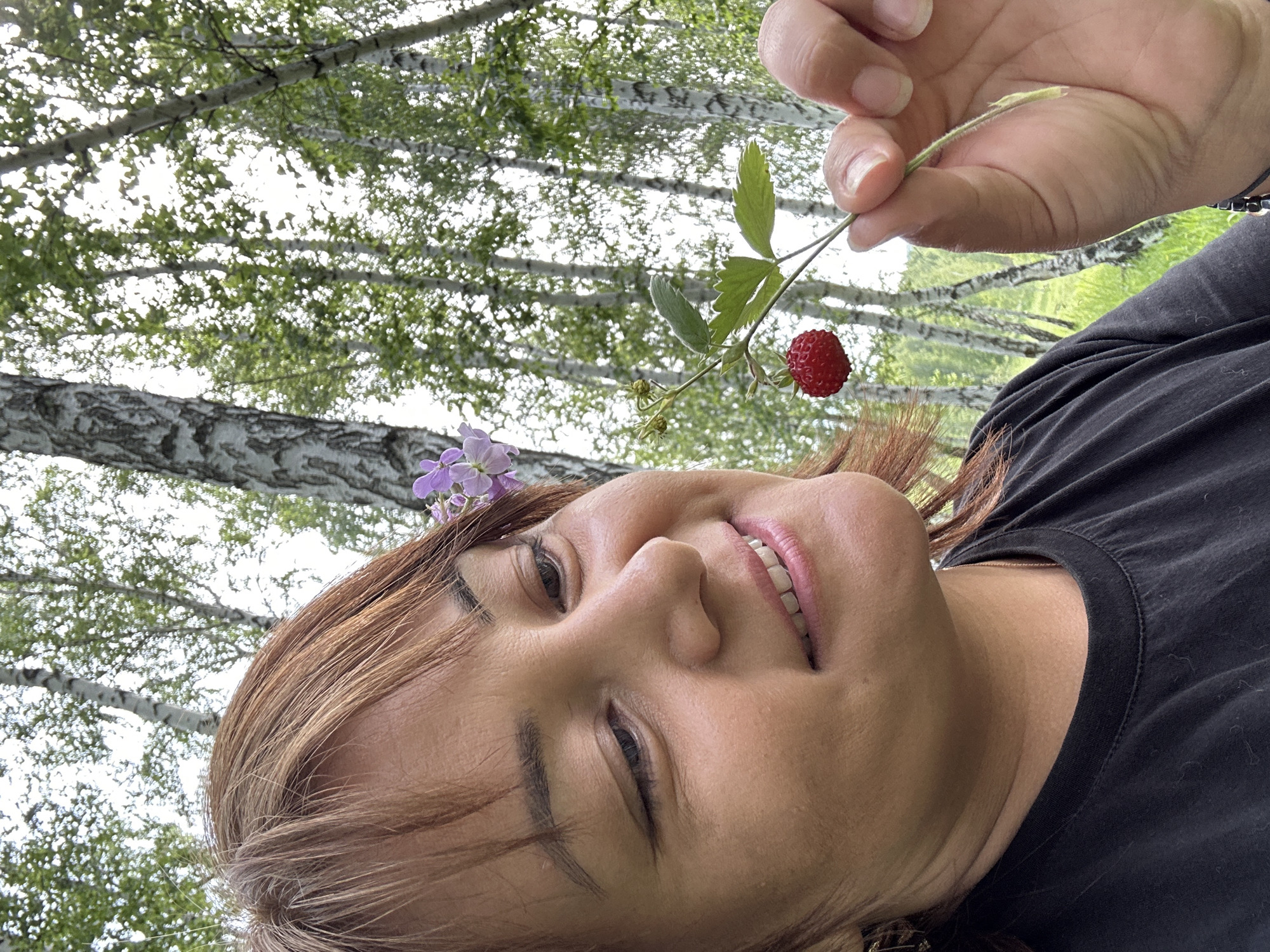 A medium shot shows a woman with auburn hair and bangs, smiling as she holds up a small red strawberry with green leaves and a purple flower near her head. She wears a black t-shirt. In the background, several birch trees with white and black bark stand tall in a grassy area under a bright sky.