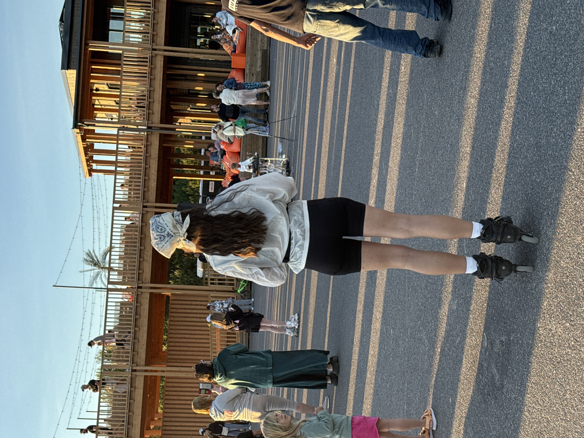 The image depicts an outdoor gathering in Tallinn, Estonia, likely during late afternoon or early evening due to the long shadows cast on the asphalt ground. In the foreground, a person wearing rollerblades, black shorts, and a white semi-sheer long-sleeved shirt with a bandana on their head is facing away from the camera. To the left, a young girl in a light green top and pink shorts stands looking towards the left. Further back, a group of people are seen milling about.
The background features a modern, multi-level wooden structure with balconies and open-air seating areas. Several people are visible on the balconies and in the seating areas, some appearing to be conversing. Orange beanbag chairs are arranged in one of the lower seating sections. Strings of lights are strung overhead between poles, suggesting a potential evening event. The overall setting appears to be a public space or entertainment venue.