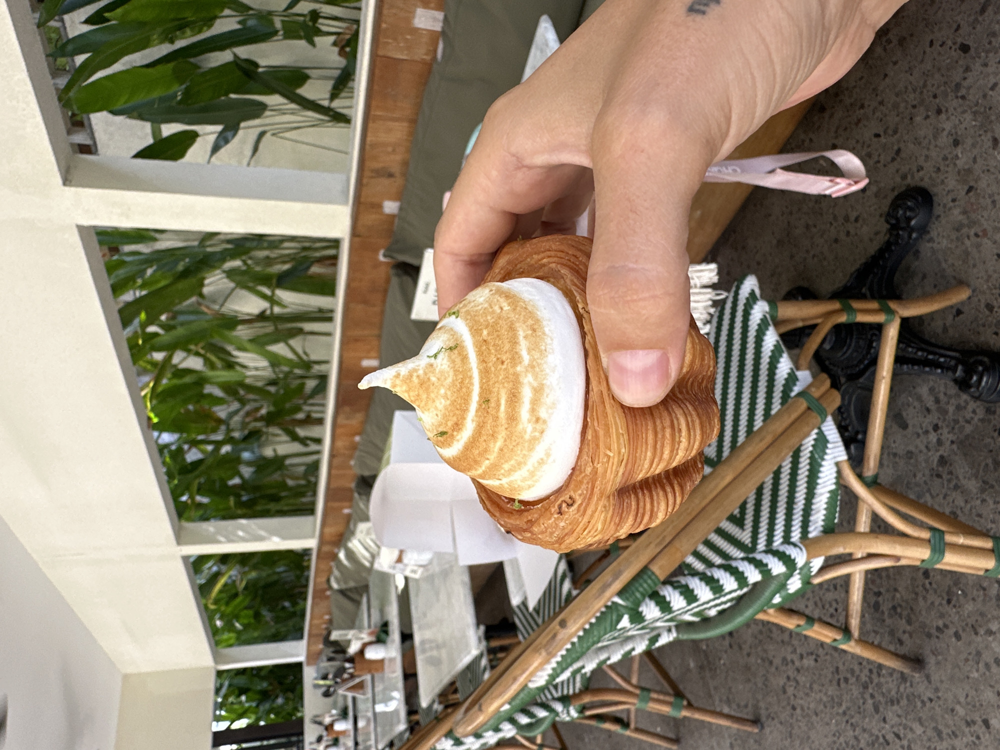 A hand holds a pastry item, a croissant with a meringue topping. The croissant's outer layers appear flaky and golden-brown. The meringue has a swirled pattern of golden brown and white. The hand is light-skinned, and a tattoo is visible on the wrist. The pastry sits atop a white paper and rests on a green and white striped cushioned chair. The chair is composed of woven materials and is part of a table set up on an outdoor patio or dining area. The patio appears to be near a building with a white trellis structure framing a view of greenery. The patio also features small tables with chairs. The environment has a casual, outdoor dining aesthetic, consistent with a cafe setting. The location is Ubud District, Indonesia.