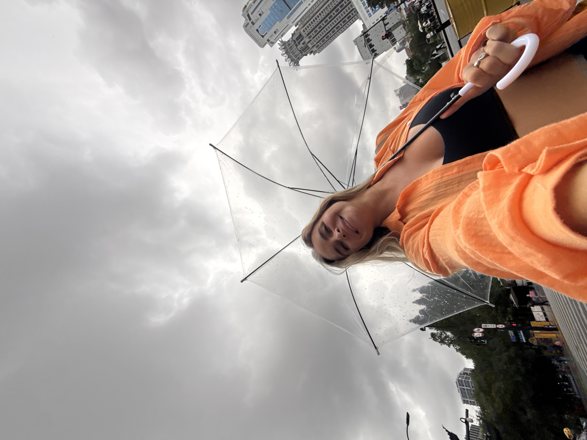 A young woman, holding a clear umbrella, stands on a pedestrian crossing on a cloudy day. She is wearing an orange open shirt over a black crop top. In the background, skyscrapers and trees are visible, suggesting an urban environment. The overall scene implies a rainy or overcast day in Shanghai, China.