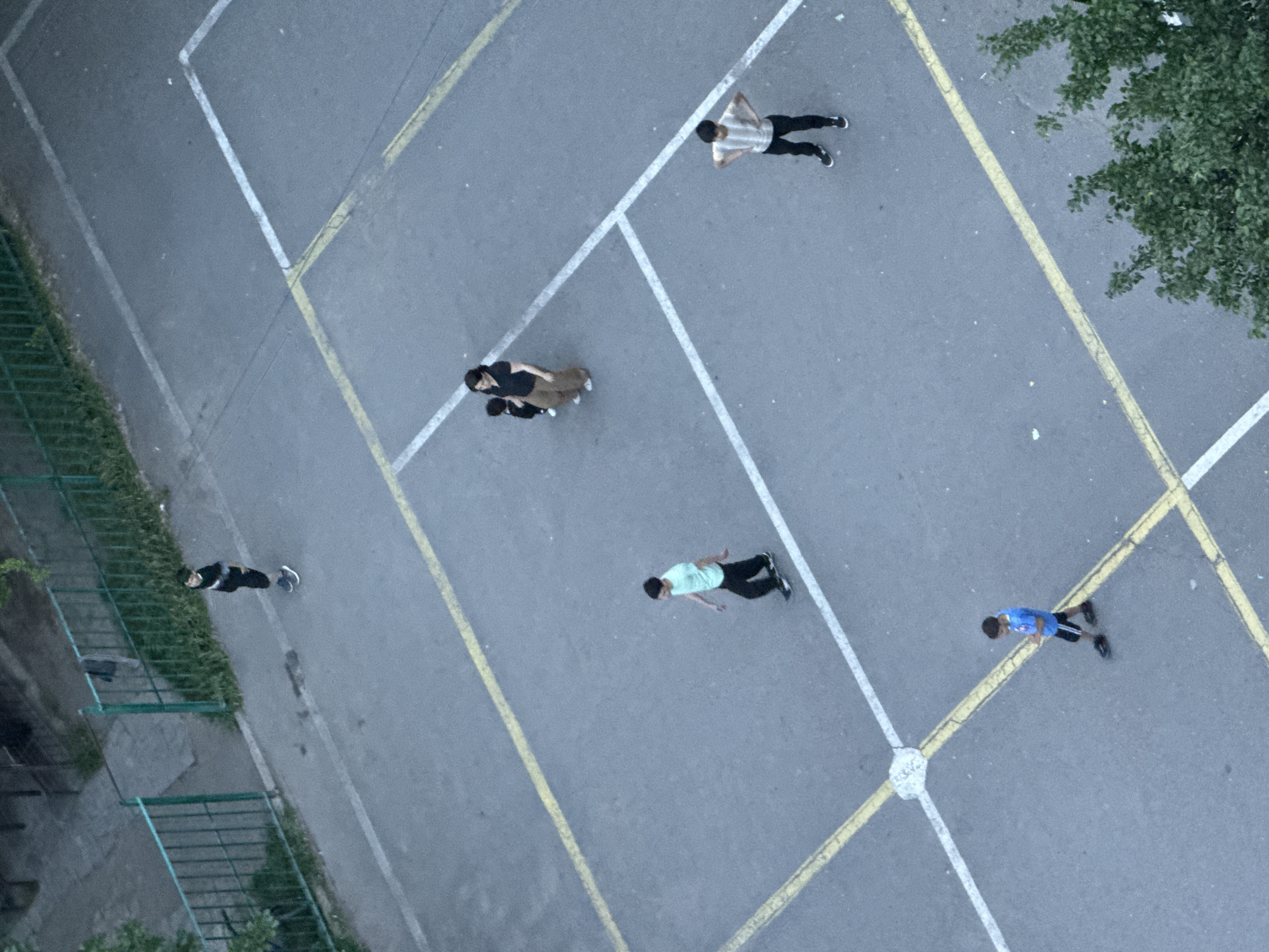 An aerial, high-angle shot captures a concrete playground with white and yellow painted lines in Abovyan, Armenia. Six individuals are visible on the playground. In the upper left, a person wearing a black t-shirt and shorts stands near a green metal fence. To the right, a woman in a black t-shirt and brown pants stands with a small child in a black outfit. Below them, a person in a light blue t-shirt and black pants walks to the left. To the right of the woman and child, a person in a striped t-shirt and black pants stands with hands on hips. In the lower left, a boy in a blue t-shirt and black shorts walks while looking down. The playground is surrounded by grass and some trees.