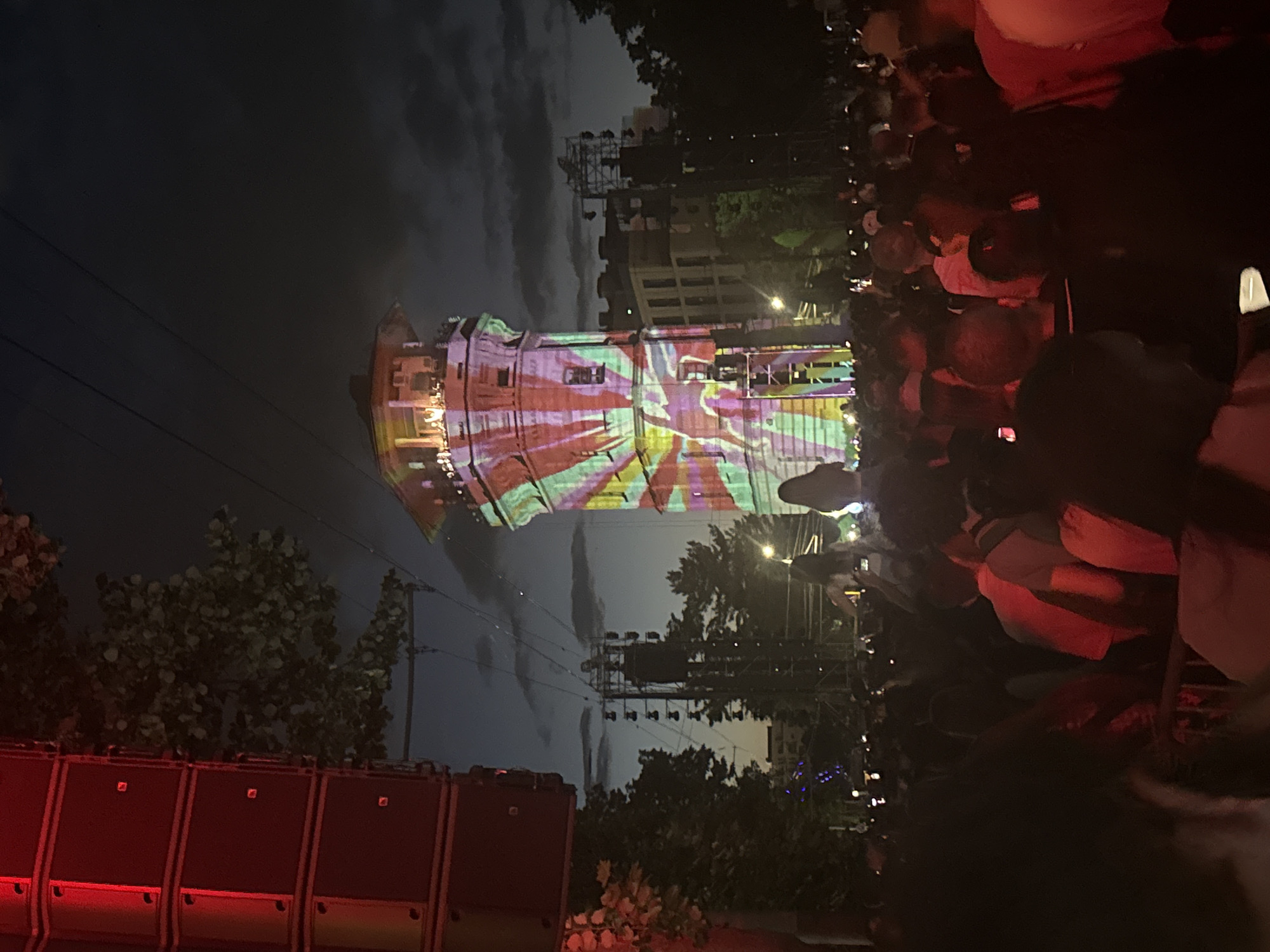 The image depicts a nighttime outdoor event in Chișinău, Moldova, near the Television Tower. A large crowd of people is gathered, facing a tower that is illuminated with colorful, abstract light projections. To the left of the frame, a large stack of black speaker cabinets is visible, with red lights emanating from some of the components. Trees and other buildings are discernible in the background. The sky is dark with some clouds, suggesting it is evening or night. The atmosphere appears to be that of a concert or festival, with the tower serving as a focal point for the visual display and likely the event's entertainment.