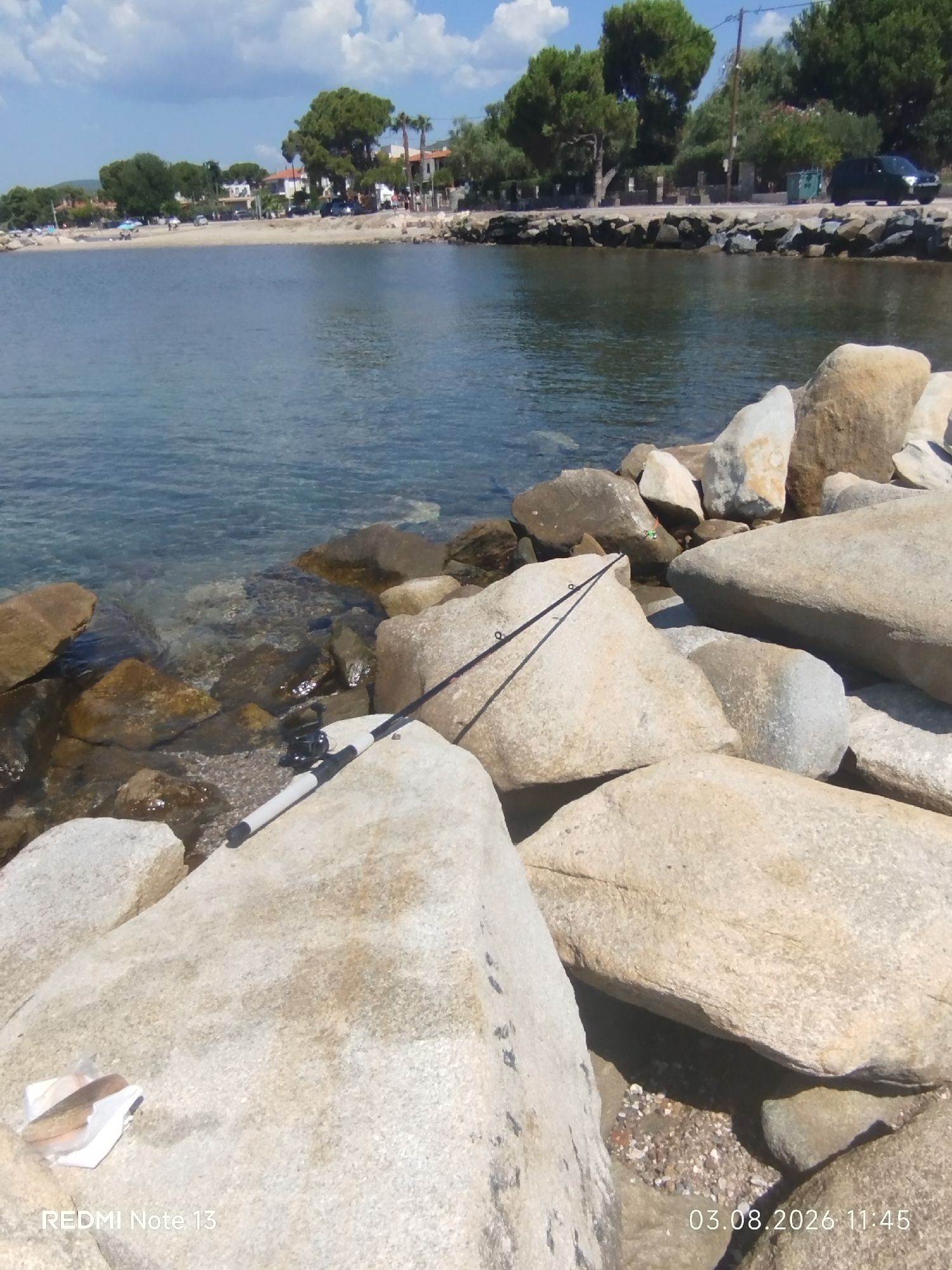 A fishing rod and reel lie on large rocks near the clear water's edge. The water is calm, and in the background, a sandy beach stretches along the coastline. Buildings and trees line the shore, with a few cars parked. The sky above is partly cloudy with a bright sun. The scene appears to be a tranquil coastal area in Sithonia Municipal Unit, Greece, possibly during a warm, sunny day.