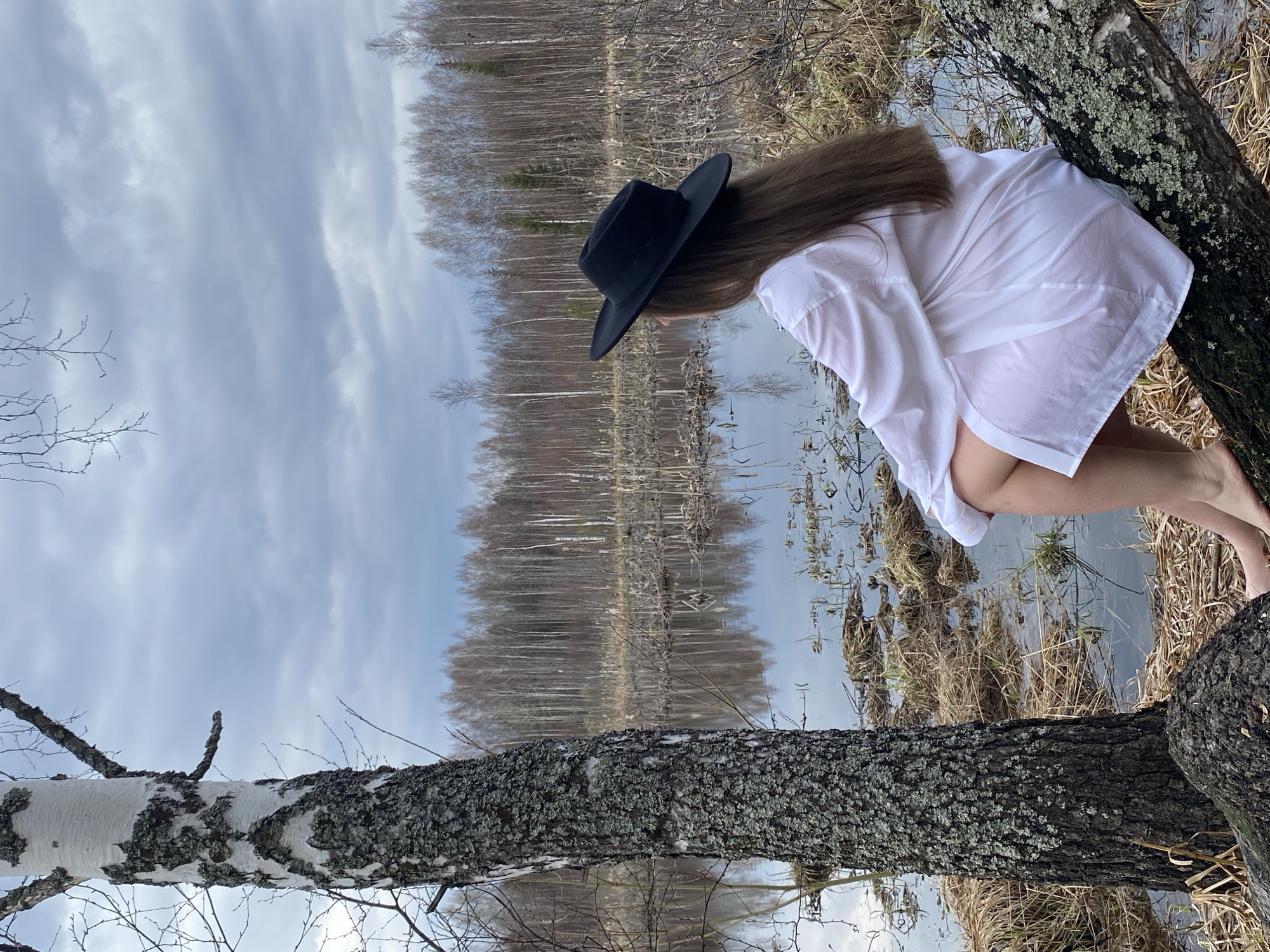 A woman wearing a white shirt and a black hat is leaning over a tree trunk by a body of water in Марковский, Russia. The water reflects the trees in the background. The sky is overcast. The woman is looking down at the water.