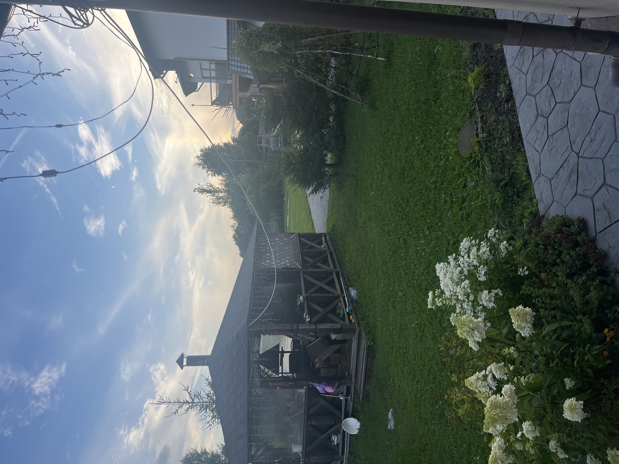 The image depicts an outdoor scene at a residential property in Vladimirovka, Russia. In the foreground, a paved patio area leads to a well-maintained lawn. A cluster of white hydrangeas and other greenery are visible in the lower left quadrant. Further into the yard, a wooden gazebo with a dark roof stands prominently. The gazebo is screened with dark netting and contains a barbecue grill and other outdoor furniture. String lights are draped along the roofline of the gazebo. Several small evergreen trees are planted in the yard, along with a decorative trellised structure covered in ivy. In the background, a multi-story house with a white facade and dark roofing is partially visible on the right side of the frame. The sky above is blue with scattered wispy clouds and a hint of a sun halo, suggesting it might be late afternoon or early evening. Electrical wires are strung across the upper portion of the image. The overall impression is of a rural or suburban backyard setting. There are no people visible in the image.