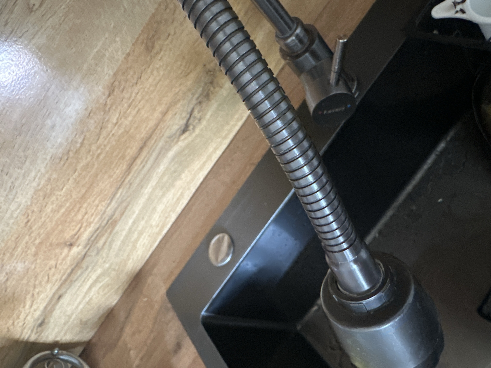 The image shows a close-up view of a kitchen sink area. A black sink is visible, along with a stainless steel faucet and its flexible hose. The faucet hose is coiled and extends from the sink. Behind the sink, a wooden countertop is visible, with a light-colored wood grain pattern. The lighting suggests an indoor setting. There are no people or animals in the image. The scene is static and shows a common household fixture. The location of Ленин айылы, Kyrgyzstan is not discernible from the image content.
