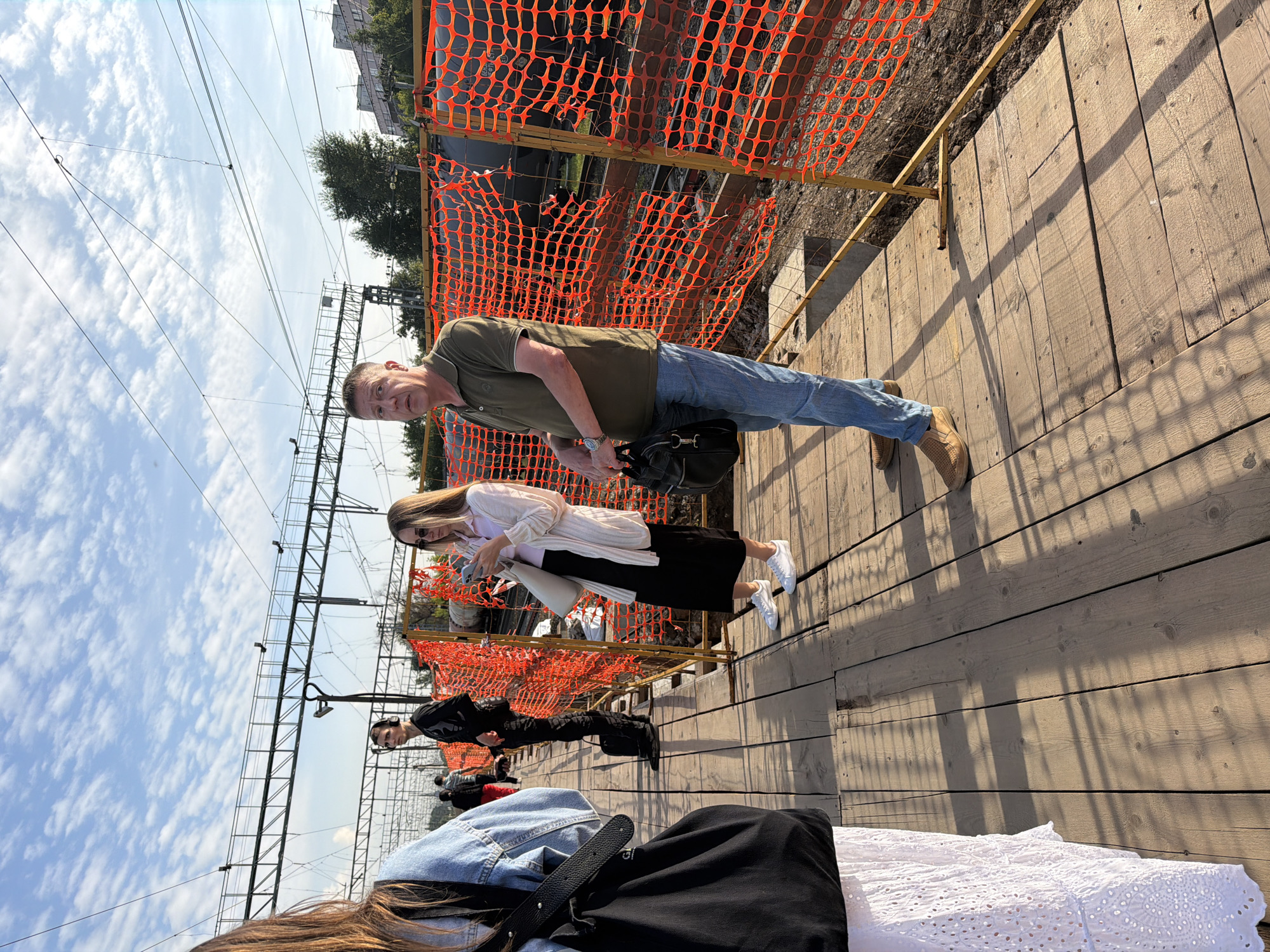 This image depicts a scene on a wooden platform, likely a train station or a similar transit area, in Shcherbinka, Russia. The platform is bordered by a bright orange safety fence, indicating ongoing construction or maintenance. Several people are visible on the platform. In the foreground, a woman is partially visible from the side, wearing a white lace skirt and a black bag. To her right, a man in a dark green polo shirt and blue jeans stands holding a black bag, looking towards the left. Further down the platform, a woman in a light-colored cardigan and black skirt is looking at her phone. Another man in dark clothing and headphones stands further back, also appearing to be waiting. The background shows train tracks, overhead wires, trees, and buildings. The sky is partly cloudy with a bright blue hue. The overall lighting suggests it is daytime, possibly late afternoon, with distinct shadows cast on the wooden planks.