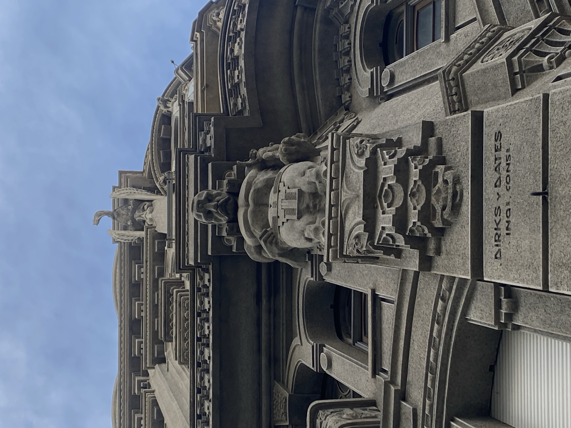 The image captures a detailed architectural view of a building facade in Buenos Aires, Argentina. The building features intricate stone carvings, including grotesque figures and decorative moldings. The name "DIRKS Y DATES, INGI. CONS." is engraved on the building. The angle of the shot is looking upwards, with a section of blue sky visible at the top. The style suggests early 20th-century architecture.