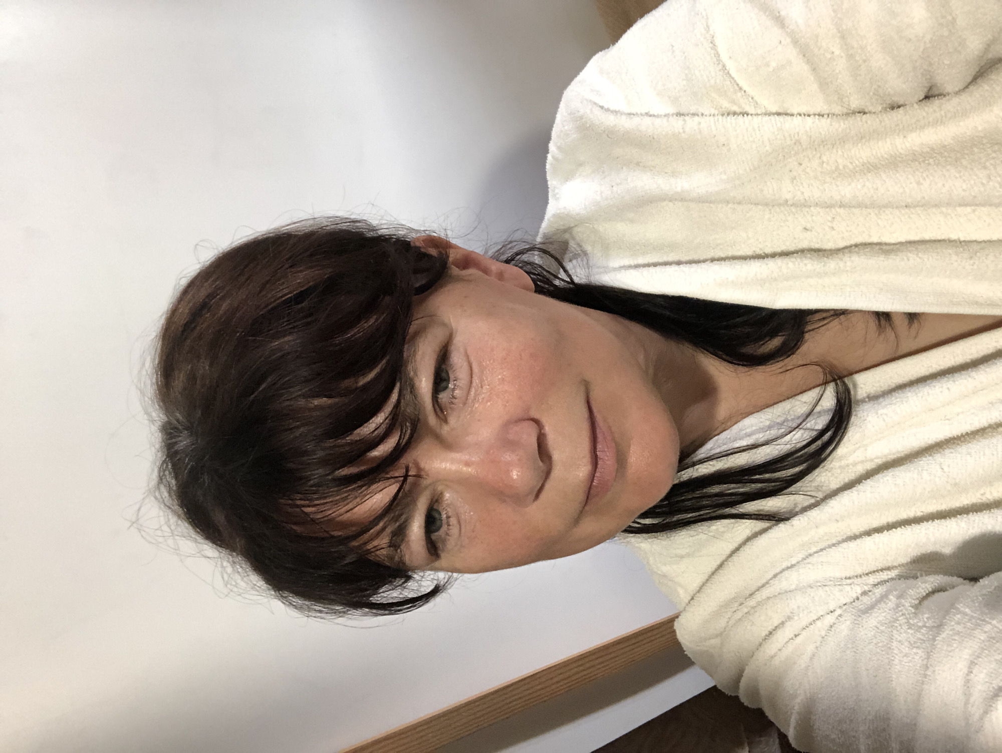 The image shows a woman wearing a white bathrobe, looking directly at the camera. She has dark brown hair and appears to be indoors, with a white wall behind her and a wooden structure to her left. The lighting suggests an indoor setting. The image is a selfie taken from a slightly high angle.