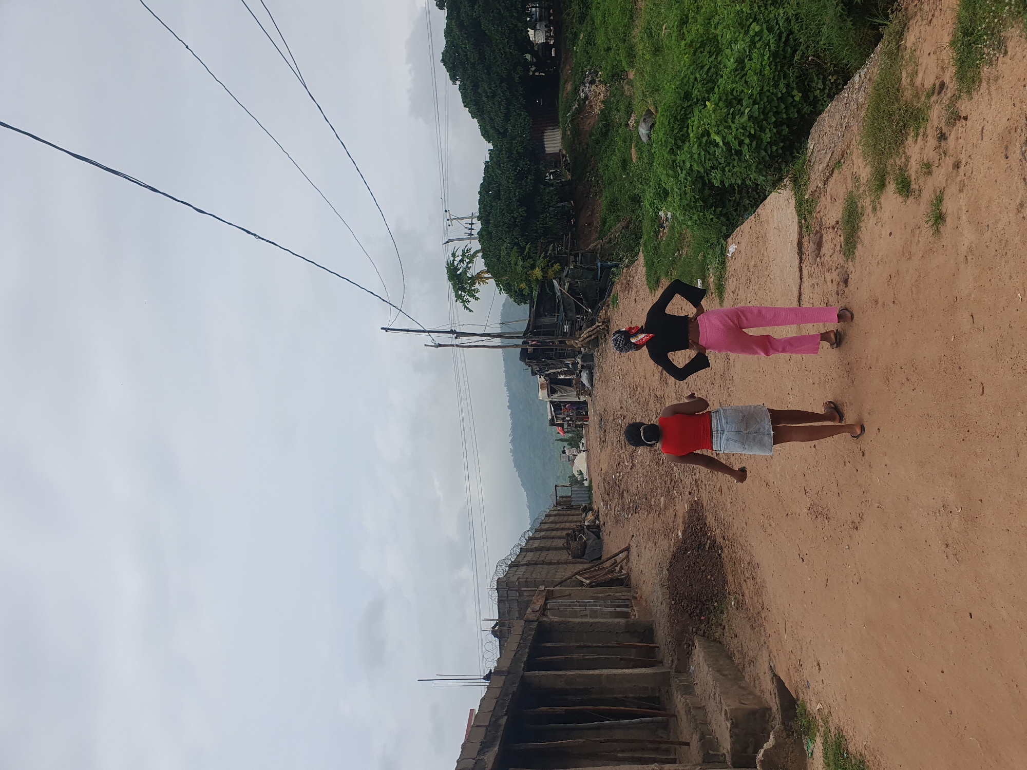 The image shows a dirt path in what appears to be a rural or semi-urban area in Lokoja, Nigeria. The path is bordered by lush green vegetation on one side and a constructed wall with barbed wire on the other. In the background, a hillside with more vegetation and a cloudy sky can be seen.
Two individuals are walking on the path. The person closer to the foreground is wearing a red top and a light blue skirt. The second person, further down the path, is wearing a black top and pink pants, with a red and white patterned headscarf. The scene appears to be during the daytime, under an overcast sky, suggesting cloudy weather.
The environment suggests a developing area, with visible construction materials and evidence of informal settlements along the path. Overhead power lines stretch across the frame, indicating the presence of electricity infrastructure. There is no visible text in the image. The overall mood is neutral, depicting everyday life in the observed location.