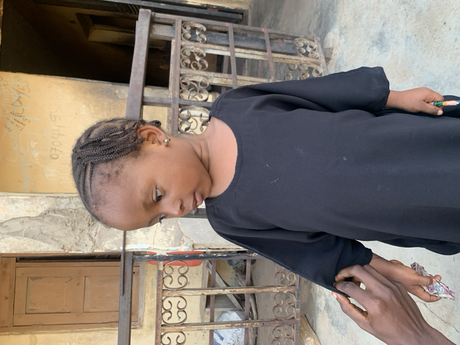 A young, dark-skinned child with dark braided hair, styled in cornrows across the top and loose braids on the sides, stands in an outdoor setting in Okene, Nigeria. The child wears a black, long-sleeved tunic with a round neckline and small green hoop earrings, gazing downwards and to the right. An adult's darker-skinned hand is visible, adjusting the left sleeve of the child's tunic. The child's right hand holds a multicolored pencil or crayon, while the left hand appears to hold a small, folded piece of patterned material. The background consists of a light brown, weathered building wall with a textured, possibly stucco finish. Faded black text and numbers, including "070449...", are visible written on the wall. A light brown wooden door is partially visible on the left side of the frame. Across the mid-ground, a dark, ornate metal railing with scrolled patterns is present, behind which some wooden debris or planks are visible. The ground beneath is a light-colored, flat surface.