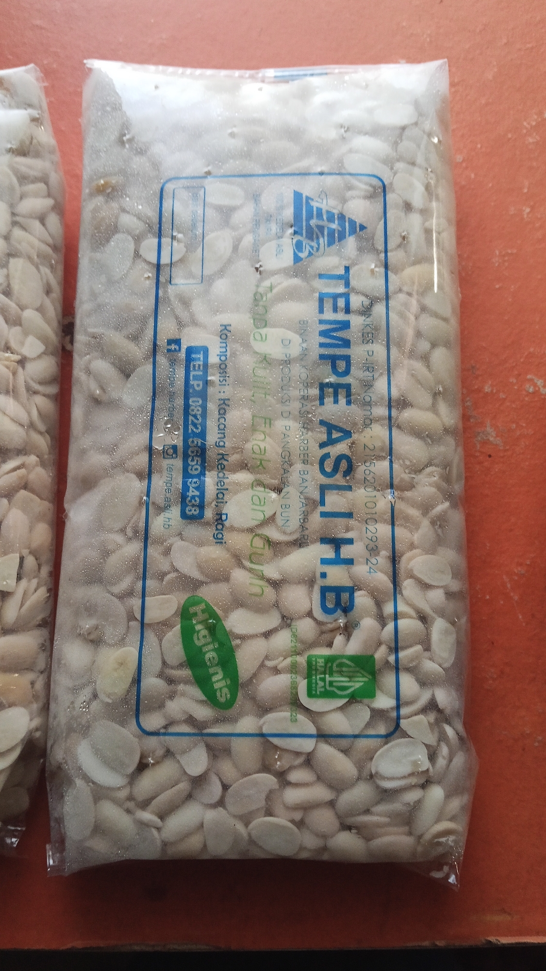 A package of tempeh, a fermented soybean product, is displayed against an orange background. The packaging is transparent, revealing the light-colored soybeans within. A blue label is affixed to the front of the package. The label contains text in Indonesian, including "TEMPE ASLI H.B.", "Komposisi : Kacang Kedelai, Ragi", and a phone number "TELP. 0822 5659 3438". There is also a logo that appears to be a stylized letter 'A' with a number '75' inside. A green oval sticker with the word "higienis" (hygienic) is visible. Another green sticker with the halal logo is also present. The image shows no people or specific landmarks of Pelanjau Jaya, Indonesia. The lighting is natural and appears to be coming from above.