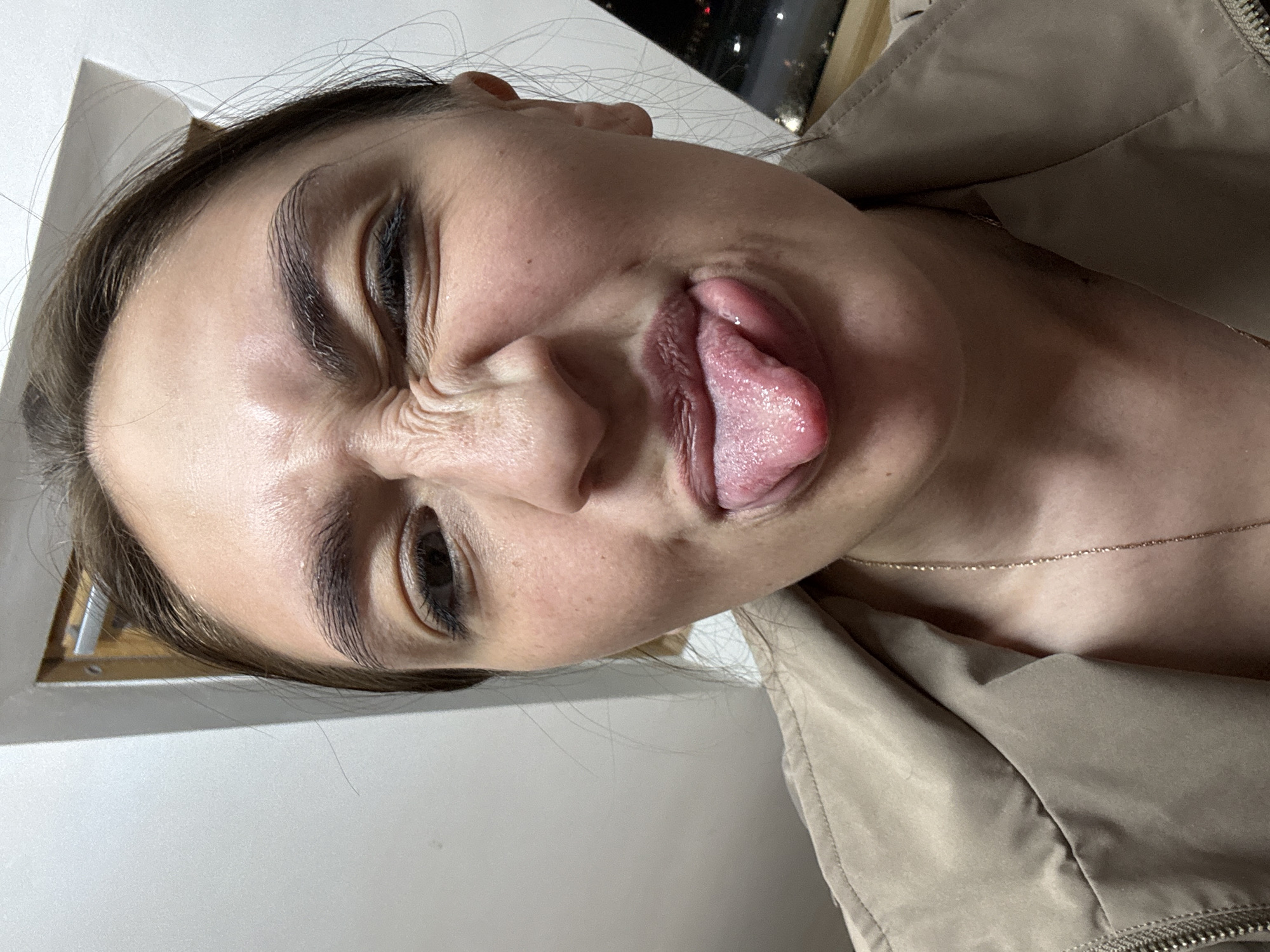 A close-up, angled shot depicts a woman making a face with her tongue sticking out. Her eyes are squinted, and her nose is crinkled. She appears to be wearing a jacket. A gold chain is visible around her neck. The background shows an interior space with part of a window, possibly showing a nighttime cityscape. There is no indication the image was taken in Sochi, Russia.