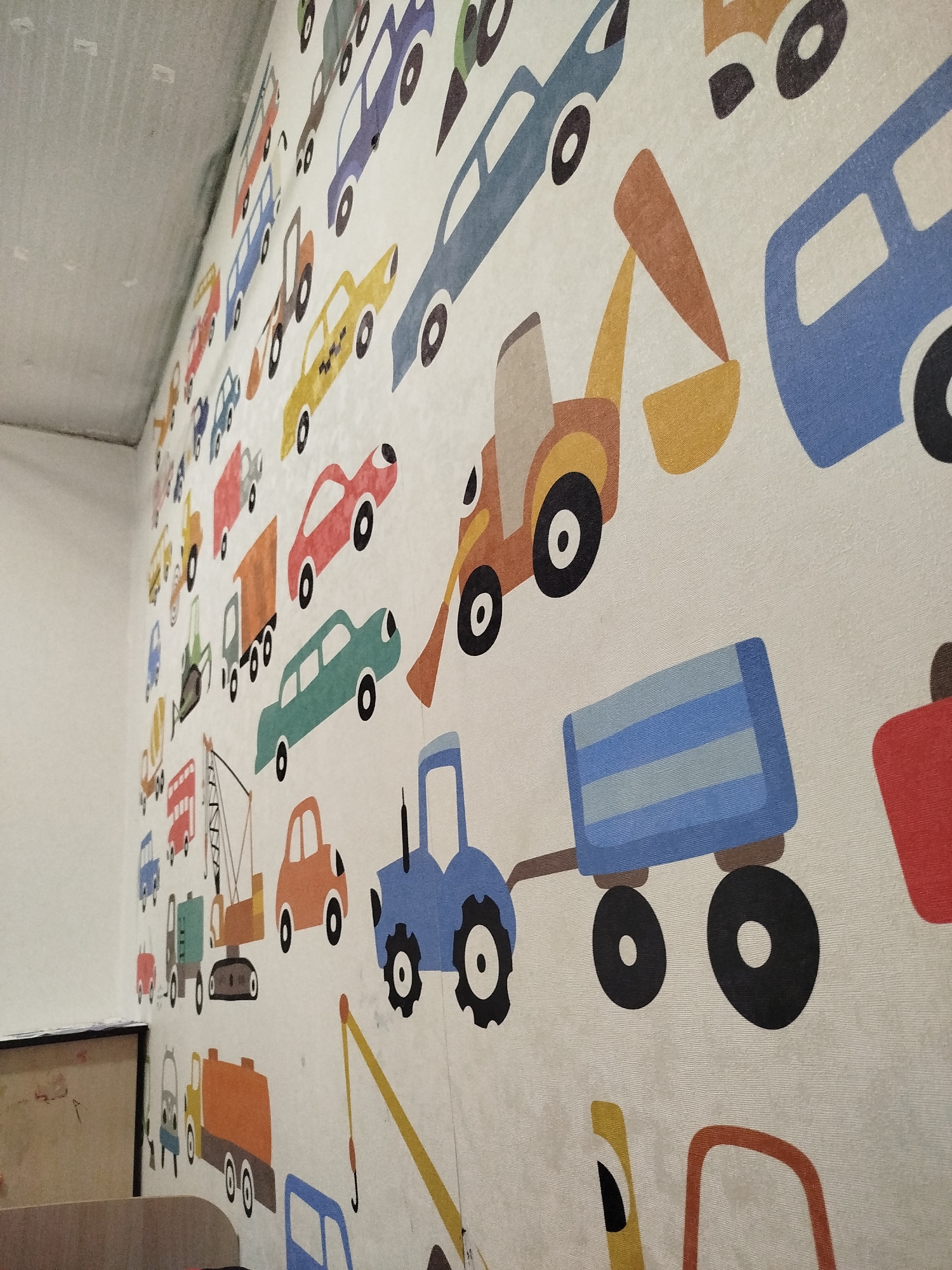 The image shows a close-up view of a wall covered with a colorful pattern of cartoon vehicles. The vehicles are depicted in a simplified, childlike style, with bold outlines and bright colors. There are various types of vehicles, including cars, trucks, buses, and construction equipment like excavators and cranes. The wall appears to be a textured, fabric-like material, and the pattern is printed or woven onto it. The lighting suggests an indoor environment, possibly a child's room or play area. The perspective is angled, looking up towards the top of the wall and to the right. The wall on the left side of the image is plain white and appears to be a standard painted wall, meeting the patterned wall at an interior corner. The scene does not provide any information that could be used to identify a specific location in Zavodoukovsk, Russia.