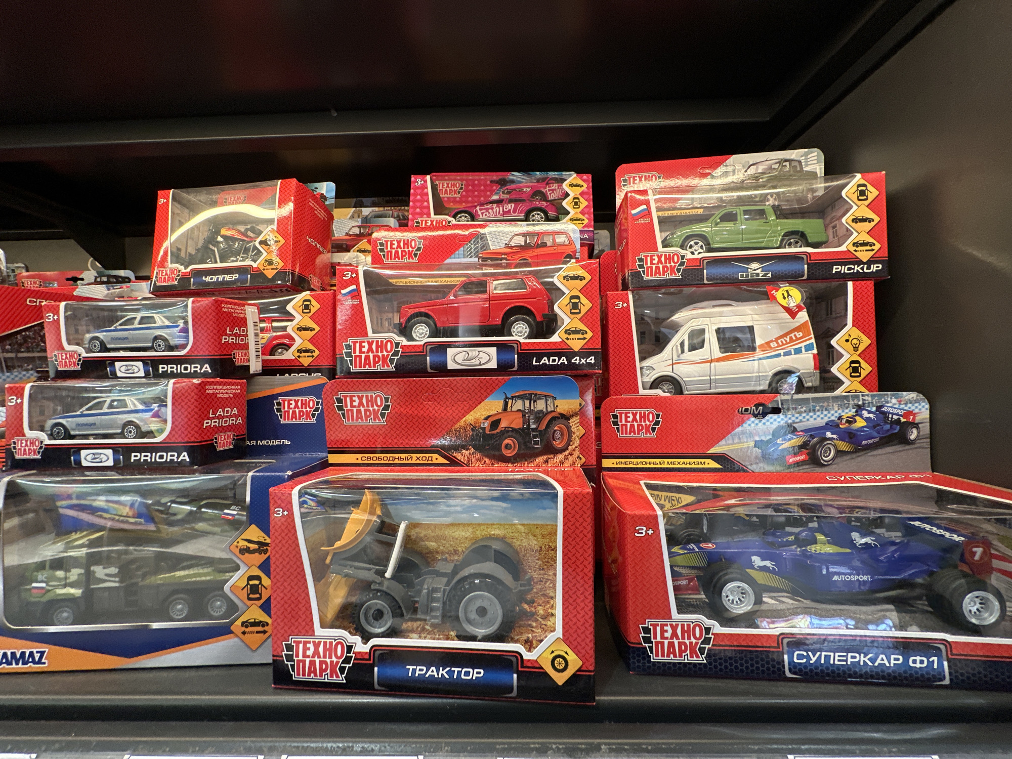 A shelf displays several "Technopark" toy vehicles in their packaging. The toys include trucks, tractors, race cars, and various car models. All packaging has "Technopark" branding and indicates they are suitable for ages 3+. Some packages have text in Russian. The scene is likely a store shelf in a retail environment but the exact location is indeterminable, including if it is in Saint Petersburg, Russia.