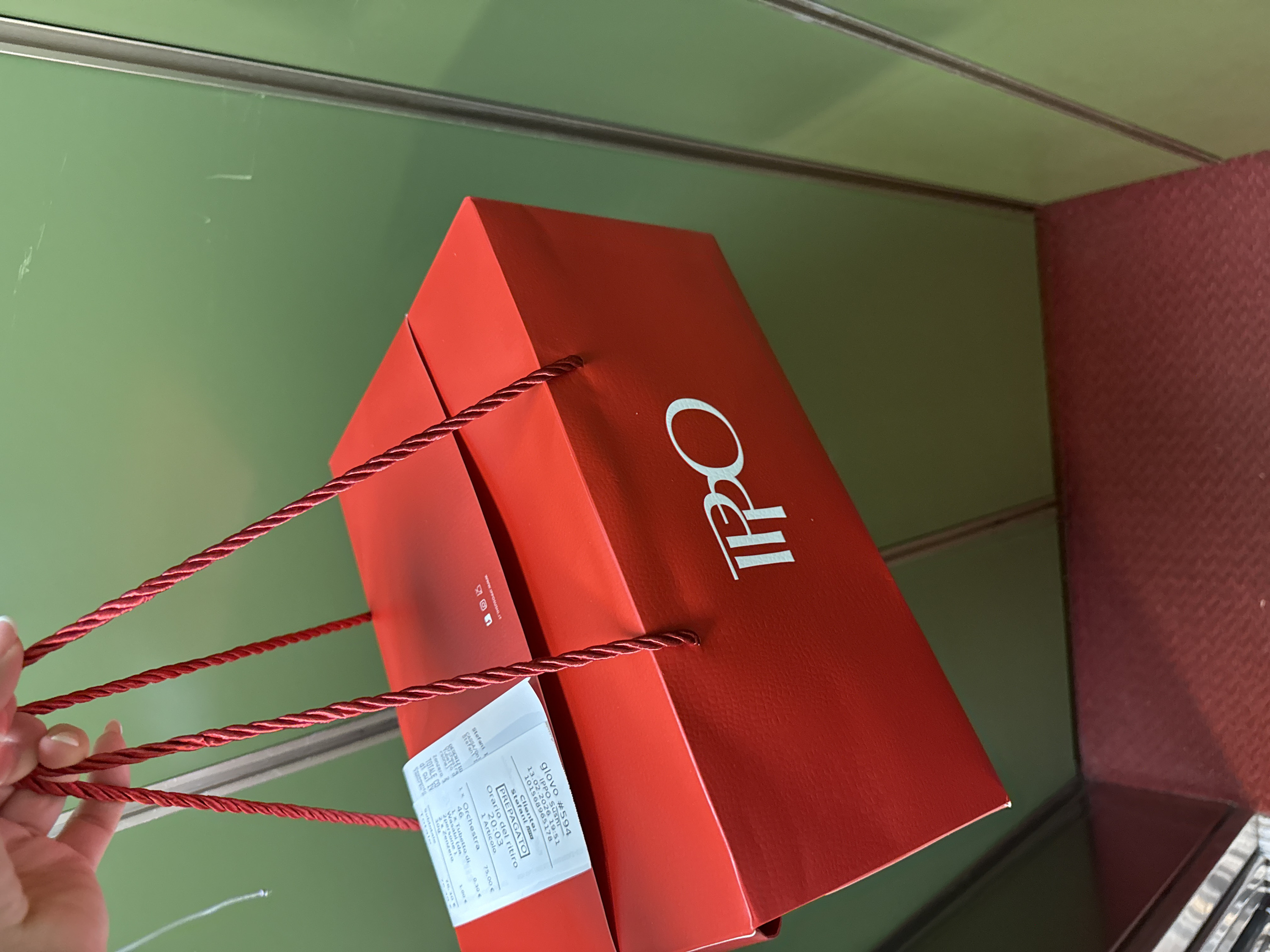 A red IPPO sushi take-out bag is held in someone's hand. A receipt is attached, displaying order #594 via Glovo for Stefani at 19:51 on February 13, 2026. The order details list "1x Orchestra" for €75.00, "Wasabi" for €0.30, and "2x Zenzero" for €1.00, resulting in a total cost of €76.30. The bag has red rope handles. The background shows a green and burgundy-colored surface.