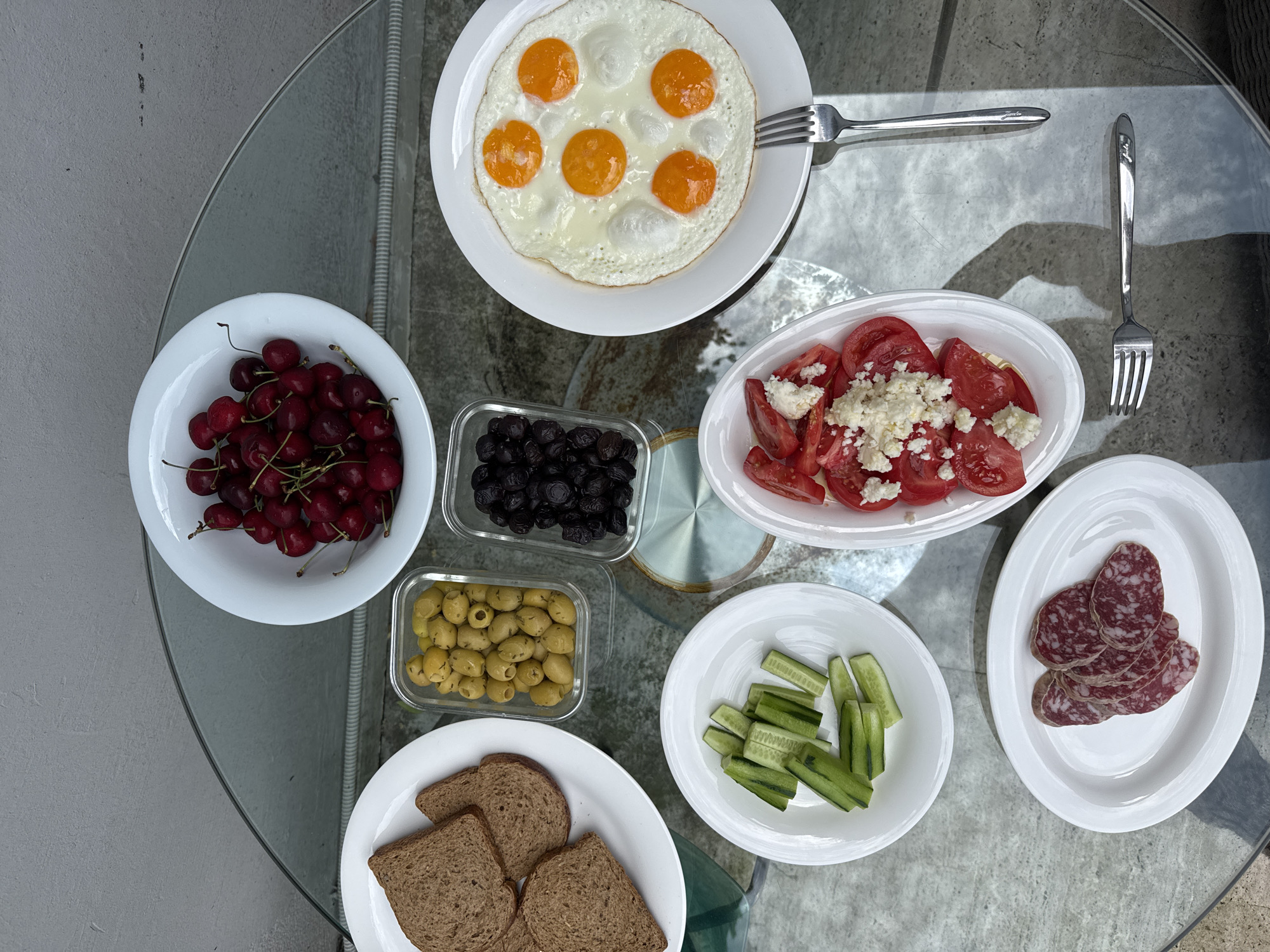 A top-down, eye-level, medium shot captures a glass-topped table laden with a breakfast spread. The scene is set outdoors, likely on a balcony or patio in Beşiktaş, Turkey, with a textured grey wall visible in the background.
Dominating the frame is a white plate of fried eggs, showcasing six distinct yolks surrounded by cooked whites. To its left, a white bowl overflows with ripe, dark red cherries. Adjacent to the cherries, a small glass container holds a cluster of green olives, and another holds black olives.
Below these, a white oval plate displays several slices of dark brown toast. Further down, a smaller white plate is filled with sliced cucumber sticks. To the right of the cucumber, a white oval dish contains sliced tomatoes, generously topped with crumbled white cheese. At the bottom of the frame, another white oval plate holds thin slices of red and white marbled salami.
Two silver forks are positioned on the table, one to the right of the fried eggs and another below the salami. The overall impression is that of a freshly prepared, healthy breakfast, ready to be enjoyed.