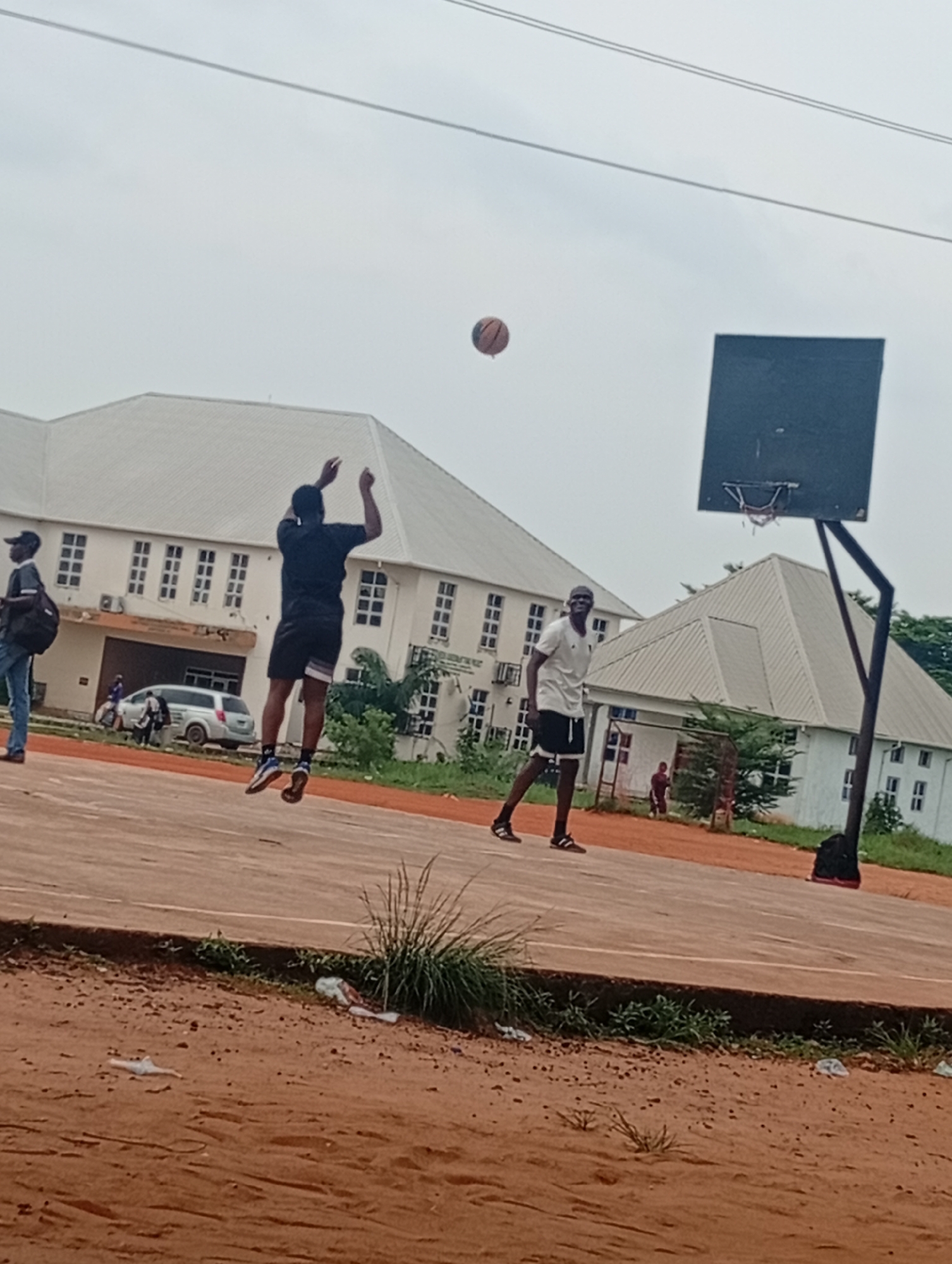 Two young men are playing basketball on an outdoor court. One man, wearing a black t-shirt and shorts, is in mid-air, reaching up to shoot a basketball. The other man, in a white t-shirt and shorts, stands near the basket, looking towards his teammate. The basketball is visible in the air above them. The court appears to be made of concrete or asphalt, with a reddish-brown surface.
In the background, a long, multi-story white building with many windows is visible, suggesting a school or university campus. A basketball hoop with a black backboard and net stands on the court. Other individuals are present in the background, including a person with a backpack standing to the left, and several people near the building and a silver van. The scene takes place outdoors, under an overcast sky, indicating a cloudy or possibly rainy day. The overall impression is a casual game of basketball, perhaps during a break or after school hours. The ground in the foreground is reddish dirt, with some dry grass and scattered white litter. The time of day appears to be daytime.