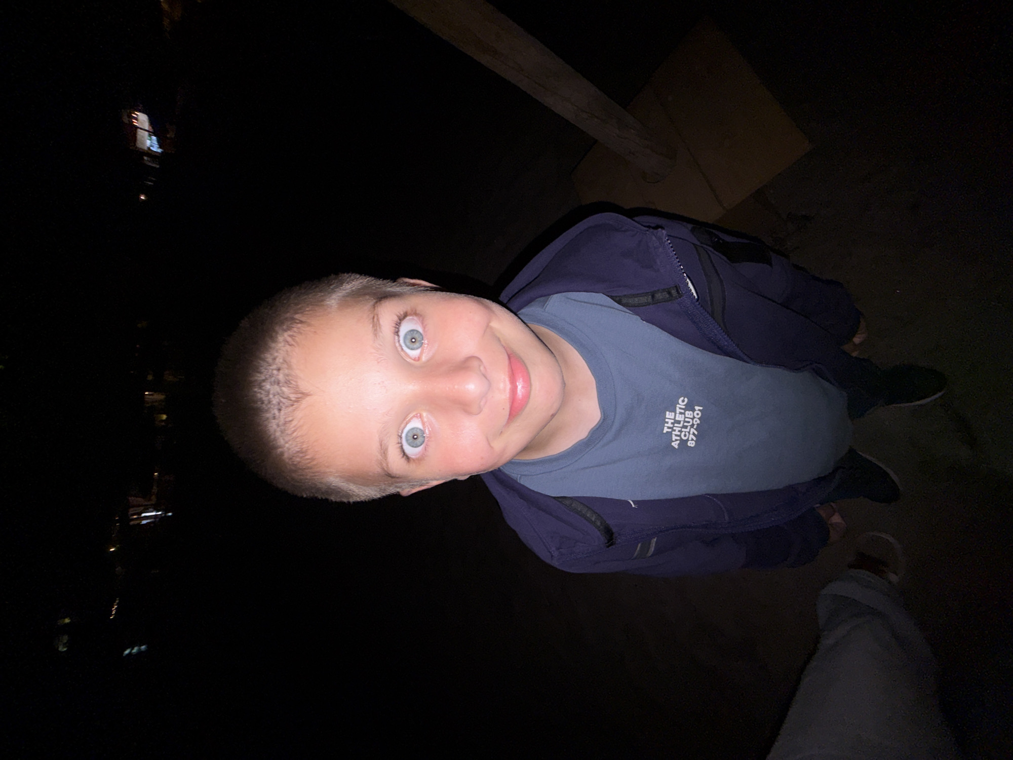 A young male, approximately 8-12 years old, is centrally positioned in an outdoor setting at night in Tsarevo, Bulgaria. He has short, light-colored hair and is looking directly at the camera with wide-open, light blue/green eyes displaying dilated pupils and a closed-mouth smile. He wears a dark blue zip-up jacket over a lighter blue t-shirt, which bears the white text "THE ATHLETIC CLUB 877-901" on its left chest. The scene is illuminated by a direct flash, creating high contrast with the extremely dark background. Distant, blurred artificial lights are discernible in the far background. To the bottom left, the lower leg and foot of another individual, wearing light-colored trousers and a light-colored sandal with a dark strap, are partially visible. Directly behind the central subject's feet, dark shoes and a glimpse of a light-colored foot or sock suggest the presence of a third individual. A rough, dark wooden beam or post is visible on the right side of the frame, extending diagonally upwards. The ground is dark and unpaved. The image is captured from a low, wide-angle perspective.