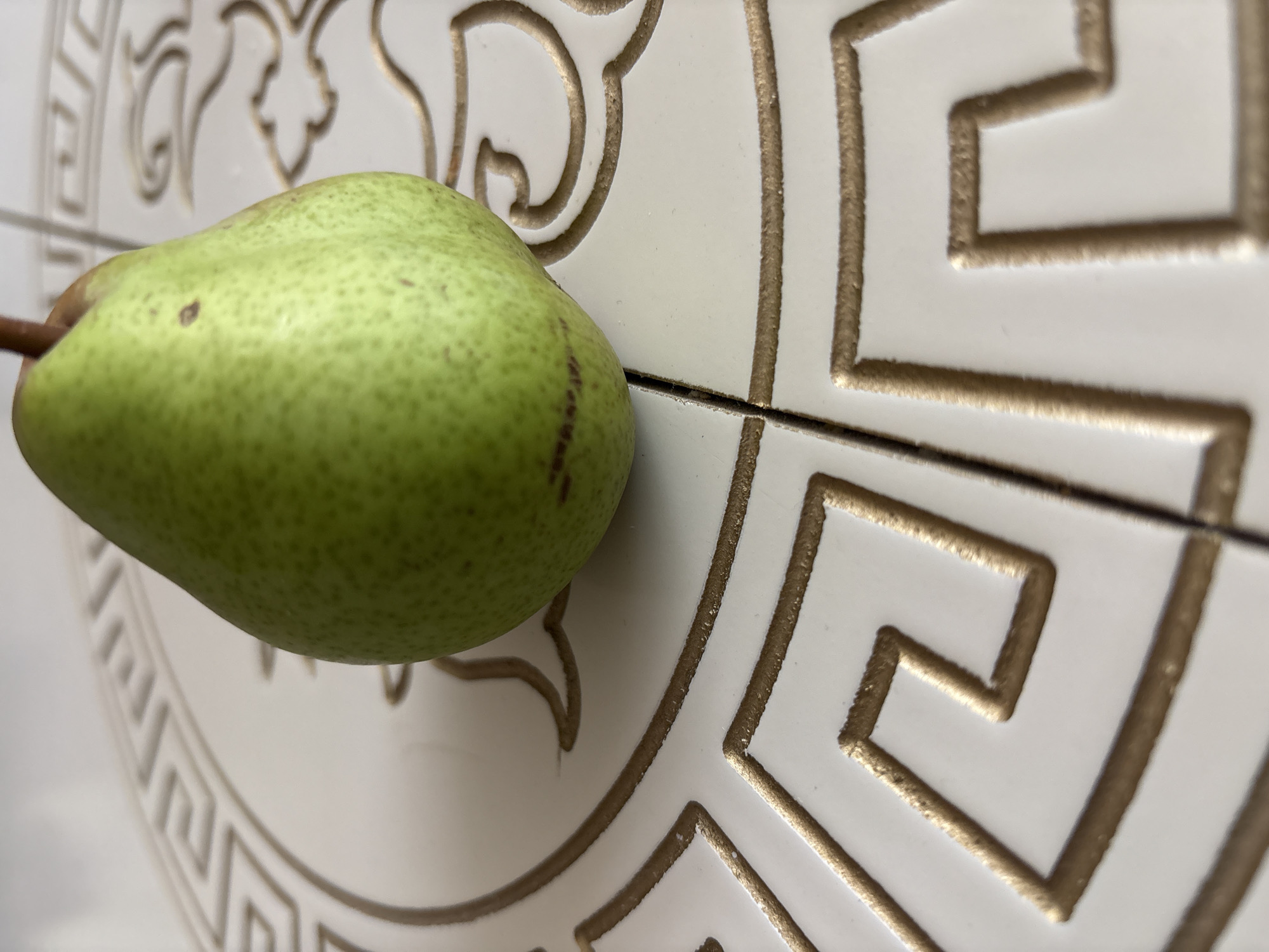 A green pear rests on a white surface with a raised gold geometric pattern. The pear is the primary subject, positioned slightly off-center to the right and towards the top of the frame. It is smooth-skinned and has a slight blush of red near its base. The background surface appears to be tiled, with a visible grout line running vertically through the lower portion of the image. The gold pattern is intricate and consists of interlocking squares and lines, resembling a meander or Greek key design. The lighting is natural and casts subtle shadows, highlighting the texture of both the pear and the decorative surface. The overall scene is static, with no visible human presence or activity. The image is a close-up shot, focusing on the detail of the pear and its immediate surroundings.