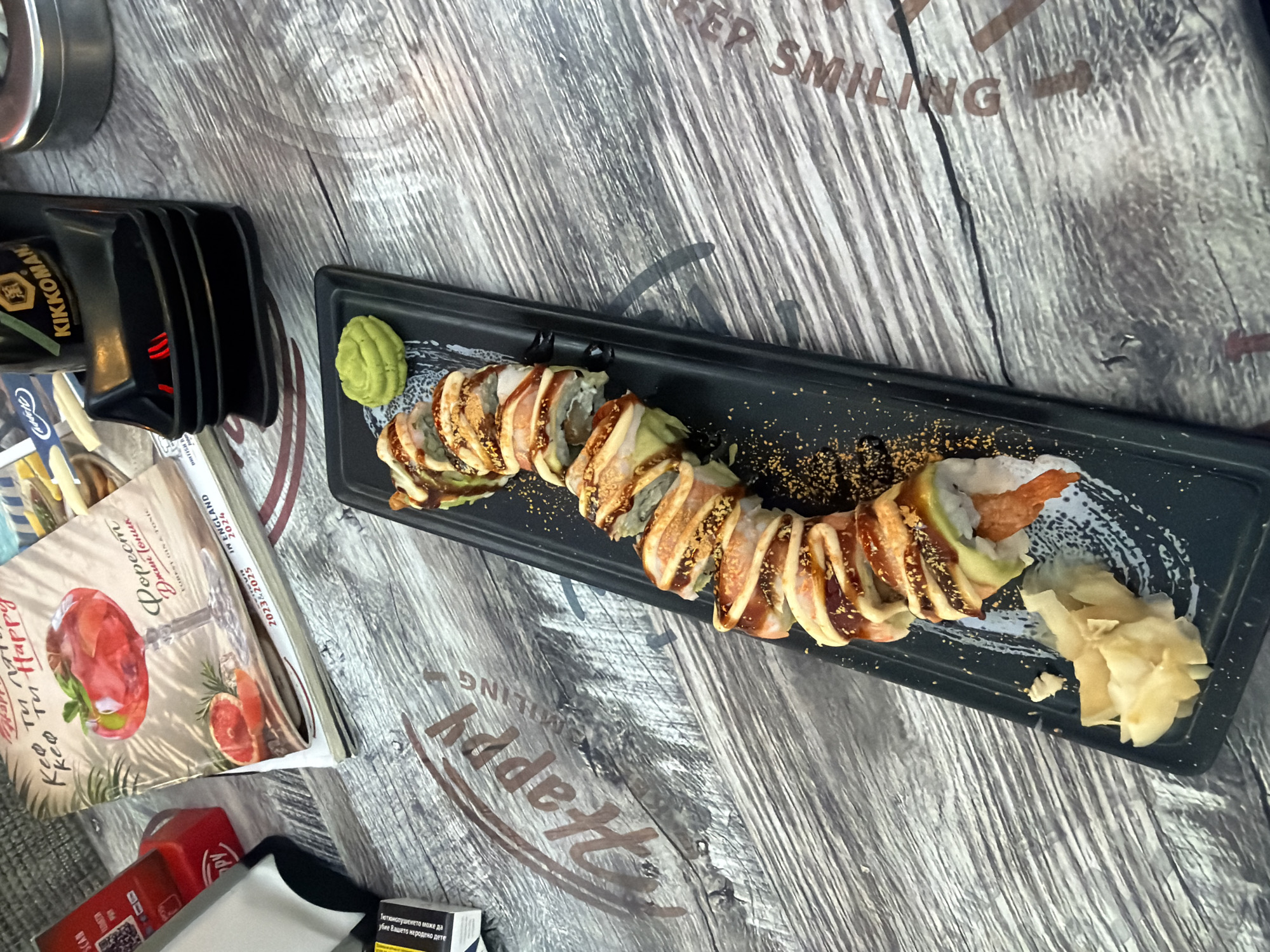 The image shows a close-up, overhead view of a plate of sushi and some accompanying items on a wooden table. The sushi is arranged in a line on a black rectangular plate, topped with a drizzle of sauce and some orange seasoning. To the left of the sushi, there are pickled ginger slices. A small mound of green wasabi sits at the top of the plate. The table surface is textured wood with a "Happy Smiling" text embossed on it. In the background, a menu with an image of a cocktail and slices of grapefruit is visible, along with a black Kikkoman soy sauce dispenser. The overall scene suggests a meal at a restaurant. The presence of the "Happy Smiling" text might indicate the restaurant's name or a branding element. Without further context, it is impossible to definitively determine the specific city or location in Burgas, Bulgaria.