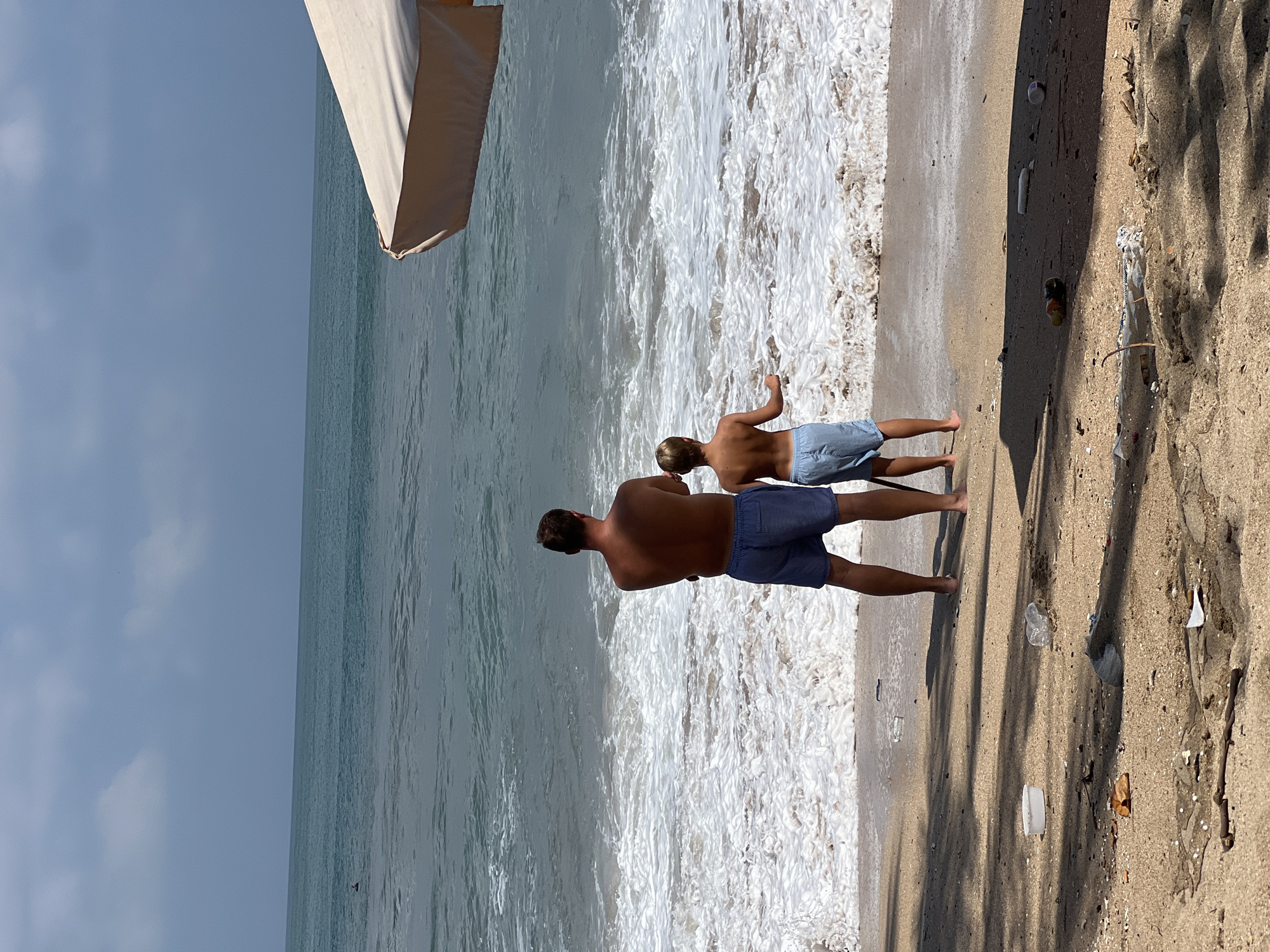 In Kuta, Indonesia, a father and son stand on a sandy beach, facing the ocean. The father, wearing blue checkered shorts, has his arms crossed. The son, in blue shorts, stands with his hands at his sides. Waves crash onto the shore, creating a foamy white line. In the foreground, the sand is littered with various pieces of trash, including plastic bottles, containers, and other debris. An umbrella casts a shadow on the sand. The sky is overcast, with patches of blue and white clouds. The ocean is a deep teal color, with ripples and waves extending towards the horizon. A single person can be seen in the distance, swimming in the ocean.