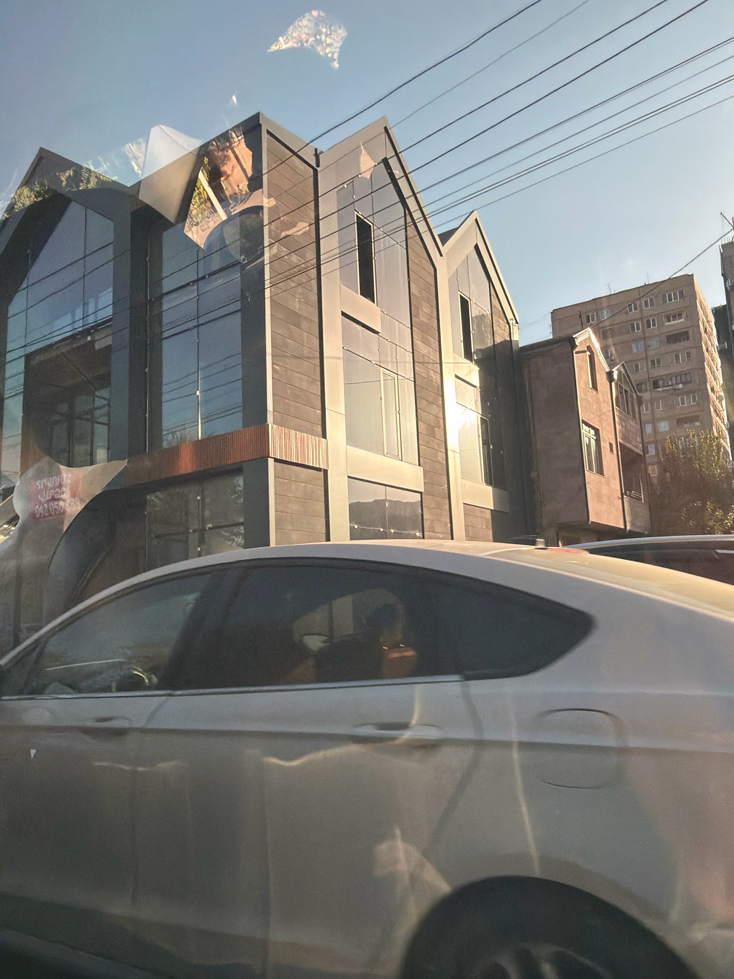 The image captures a street scene in Yerevan, Armenia, featuring a modern building with a unique, angular architectural design. The building's facade is a mix of dark, textured material and large glass windows that reflect the sky and surrounding environment. The geometric roofline is distinctive, with several pointed sections. Next to this building, a taller, older apartment building with numerous windows is visible, suggesting a mix of architectural styles in the urban landscape. The foreground is dominated by the side of a light-colored car, likely moving or parked, with reflections visible on its surface. Power lines crisscross the upper part of the image against a clear sky, indicating an urban setting. The overall impression is of a bright, possibly sunny day in a city with a blend of contemporary and older structures.