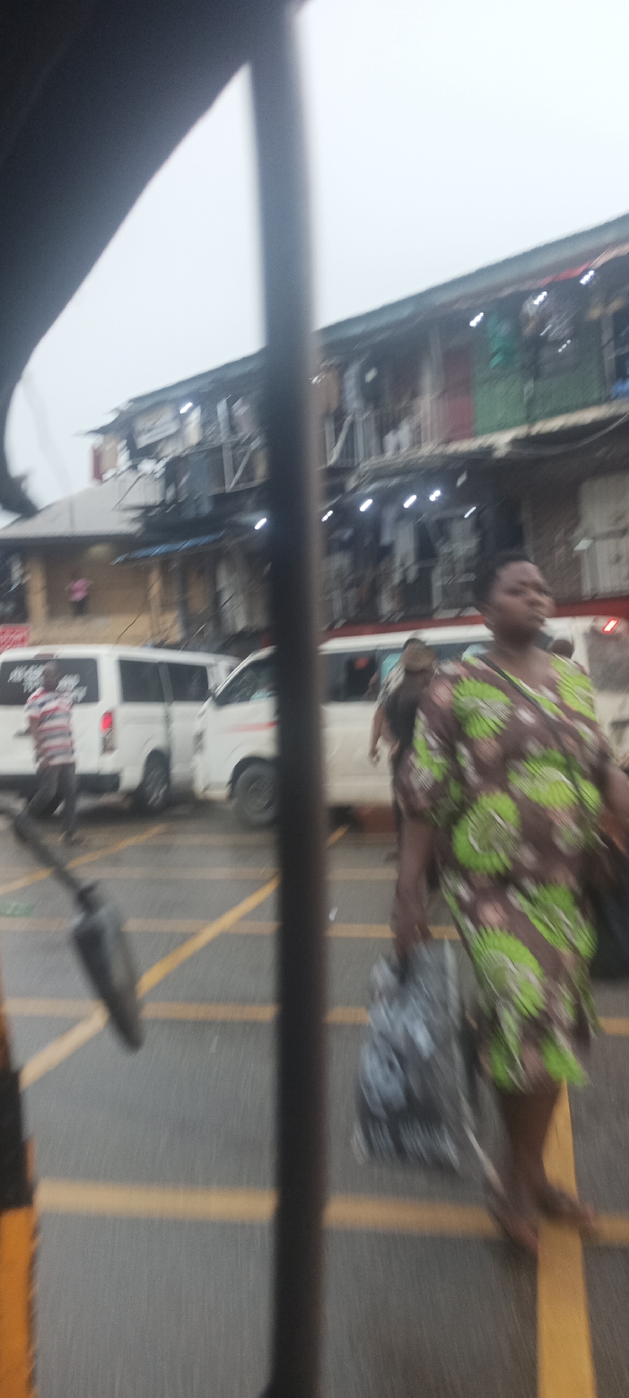 The scene is a street in Umuahia, Nigeria, appearing to be a market area with buildings in the background. The street has yellow lane markings. There are white vans parked along the side of the road. A woman in a green and brown patterned dress is walking barefoot across the street, carrying a large bag. The view is obscured by what seems to be a pole or part of the vehicle from which the photograph was taken. A man in a striped shirt is walking near one of the vans.